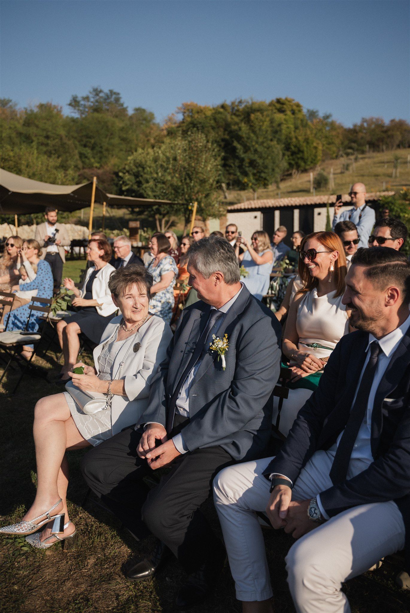 Wedding guests seated in formal attire at an outdoor ceremony with blue sky backdrop.