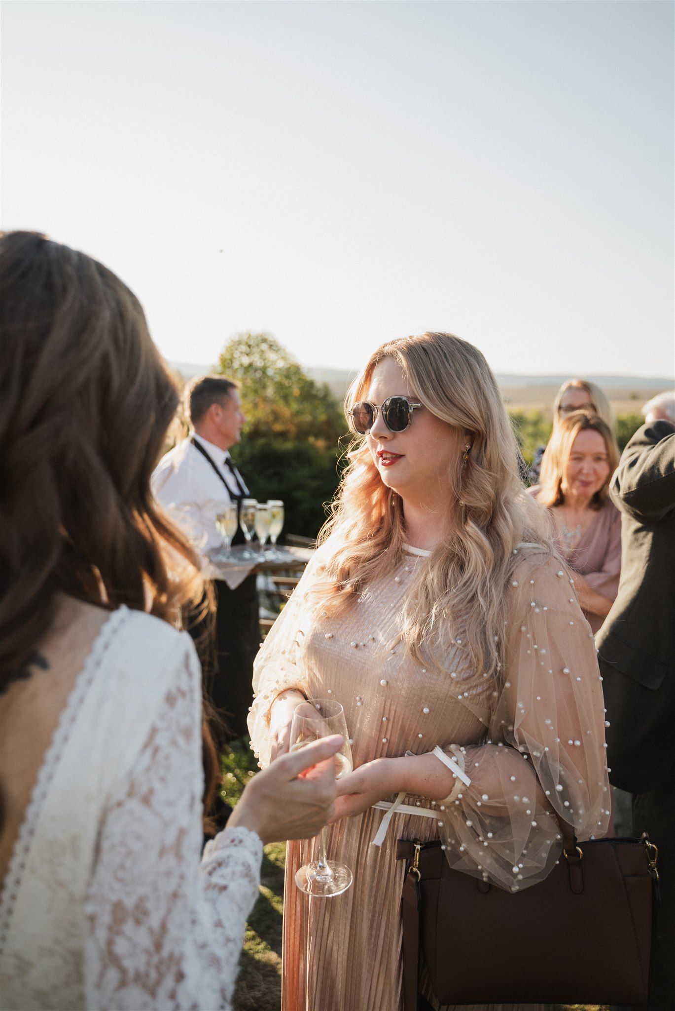 Wedding guests in elegant attire socialize during outdoor cocktail hour at sunset.