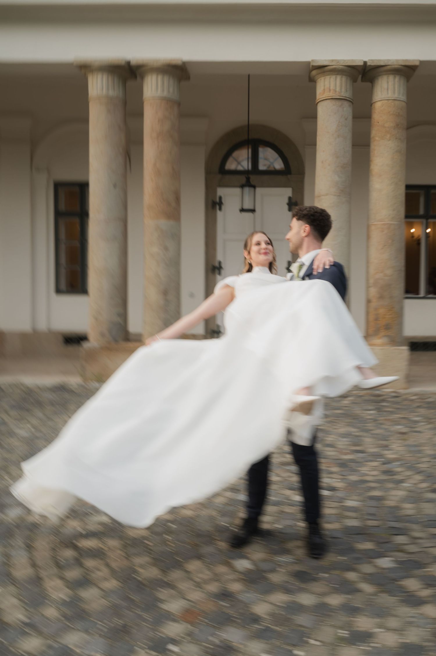 Groom spinning bride in white wedding dress outside elegant building with stone columns on cobblestone courtyard.