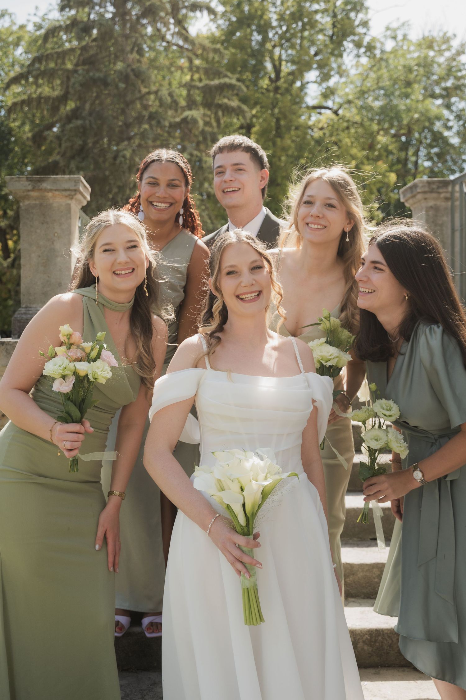 Bride in white off-shoulder gown holds calla lilies bouquet surrounded by smiling bridesmaids in sage green dresses outdoors.