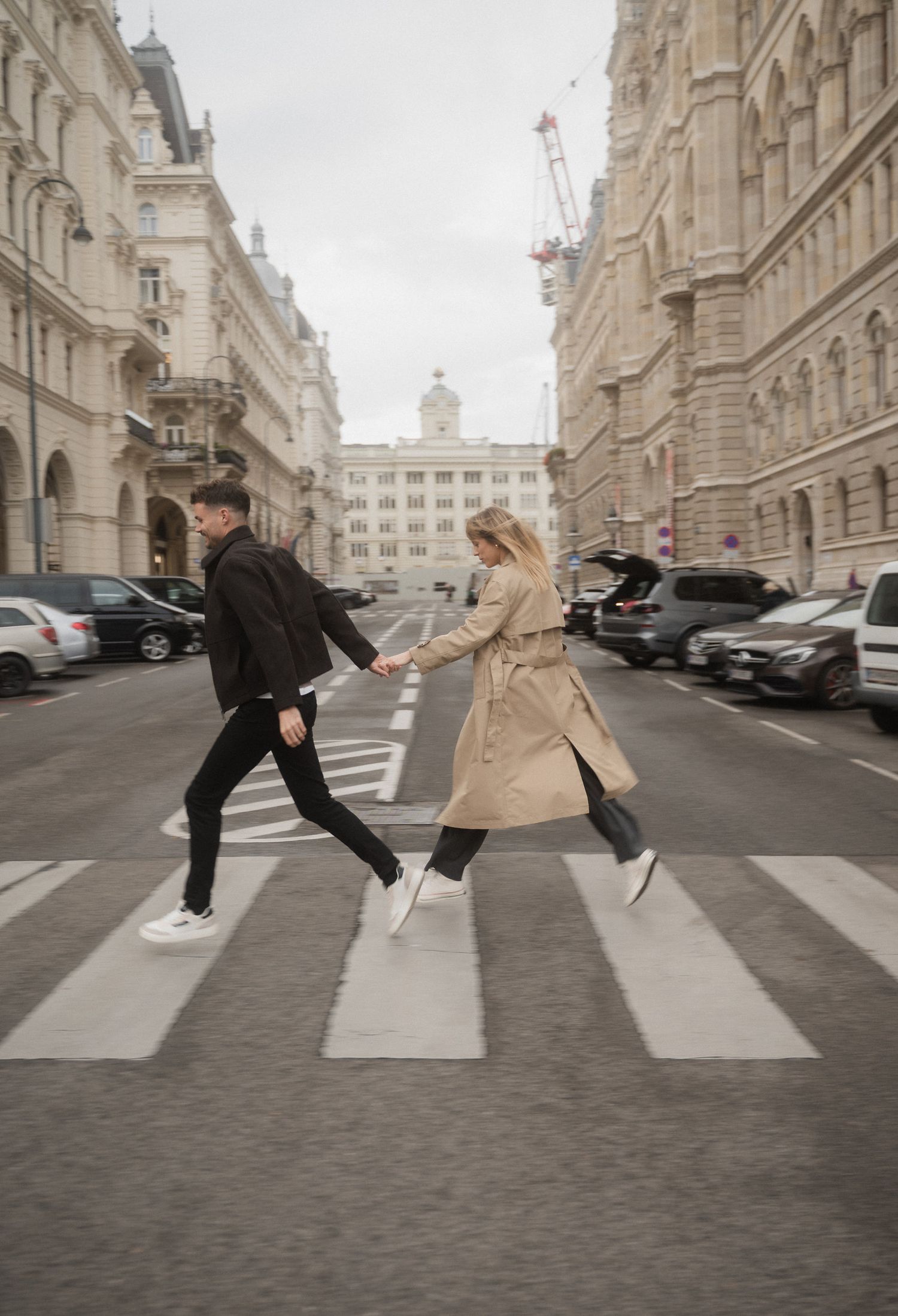Two people recreate the iconic Beatles Abbey Road album cover pose on a cobblestone street in a European city.