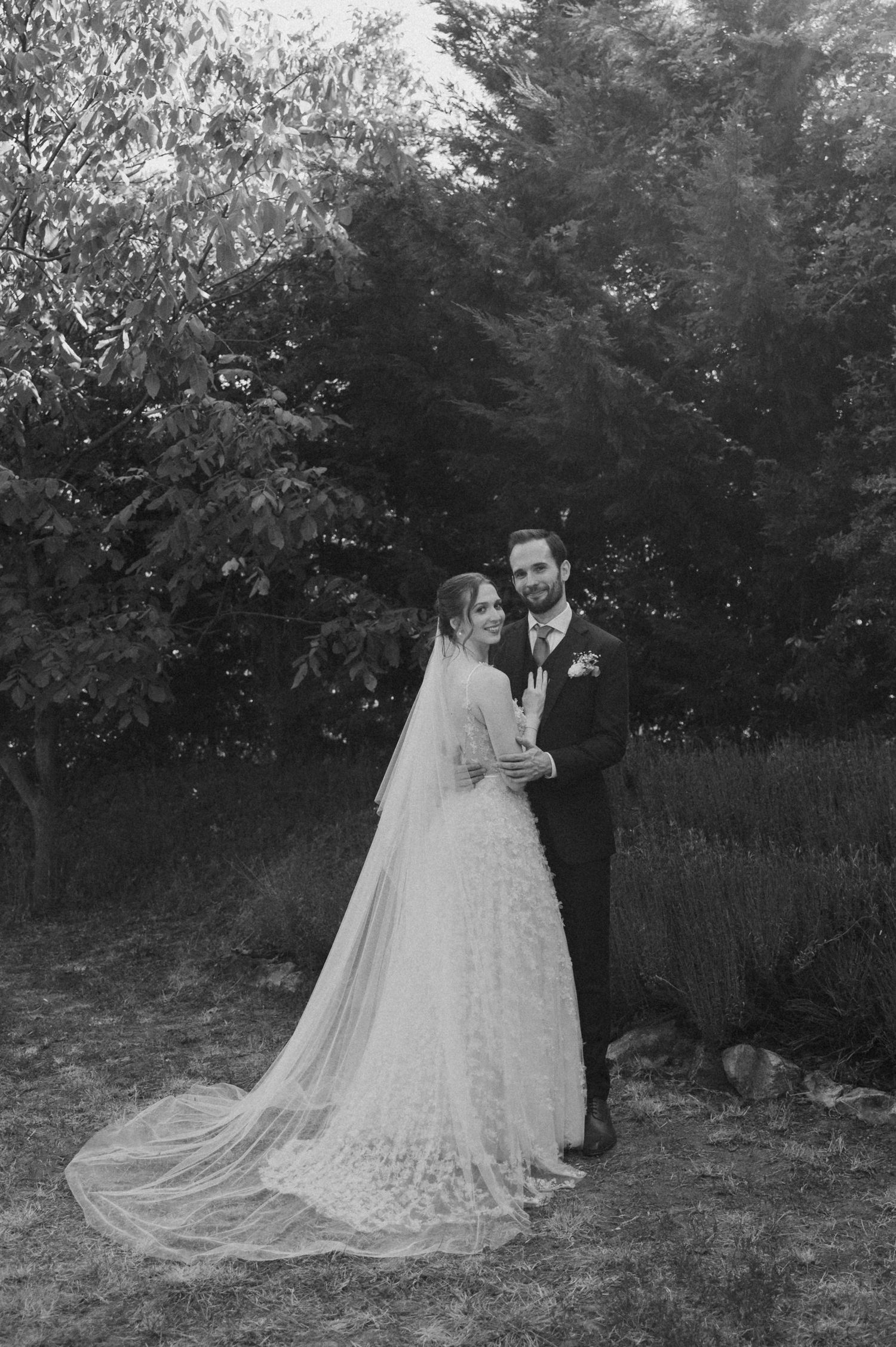 A couple in wedding attire embraces outdoors in a black and white photo with a long flowing veil trailing behind them.