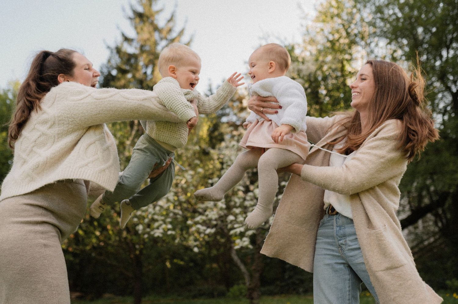 Two women playing with babies outdoors in a garden, lifting infants joyfully among green trees in spring.