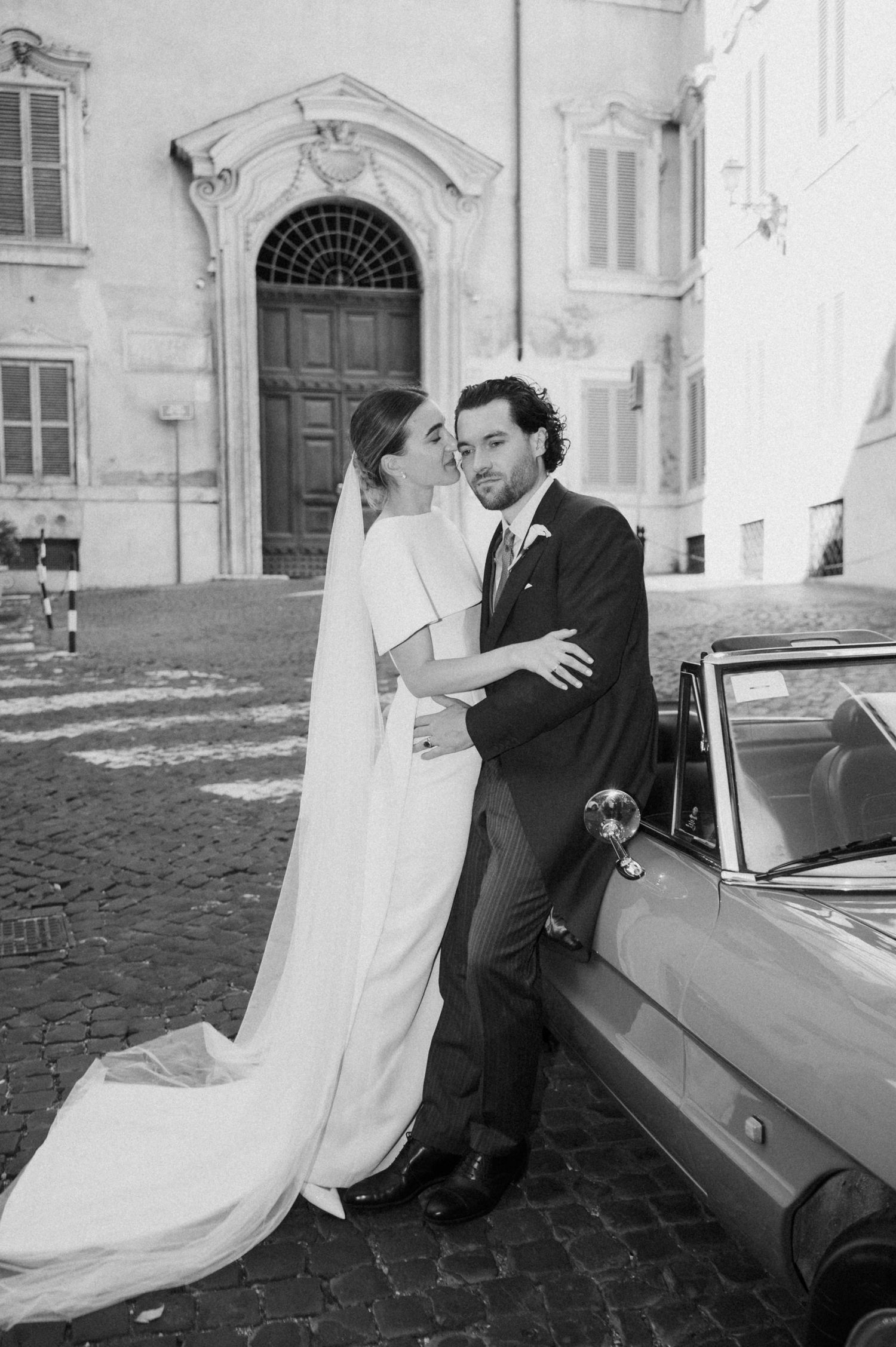 A classic black and white wedding portrait with a vintage car on cobblestone streets.