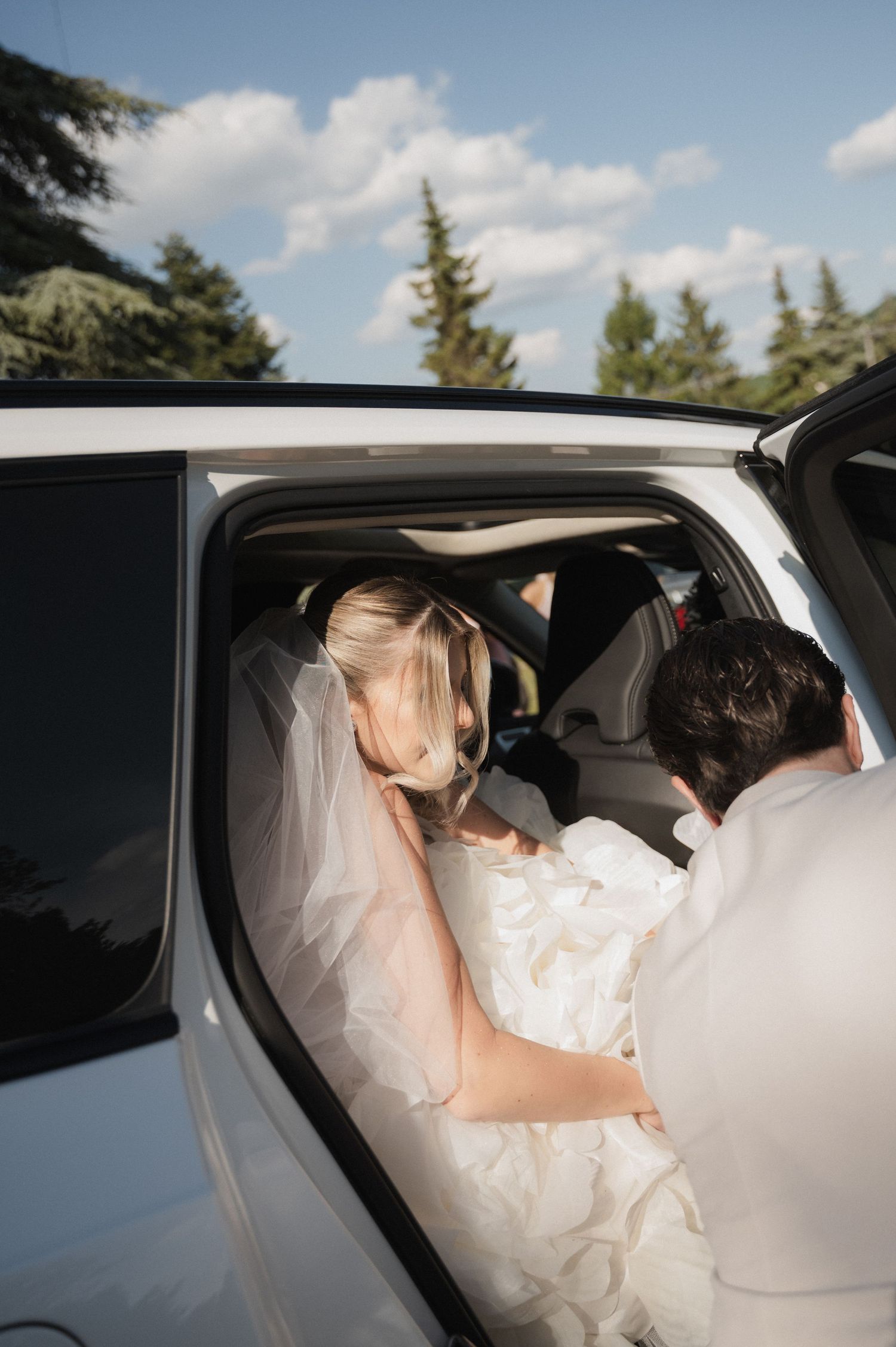 Bride in white gown and veil being helped into a white limousine by the groom on their wedding day.