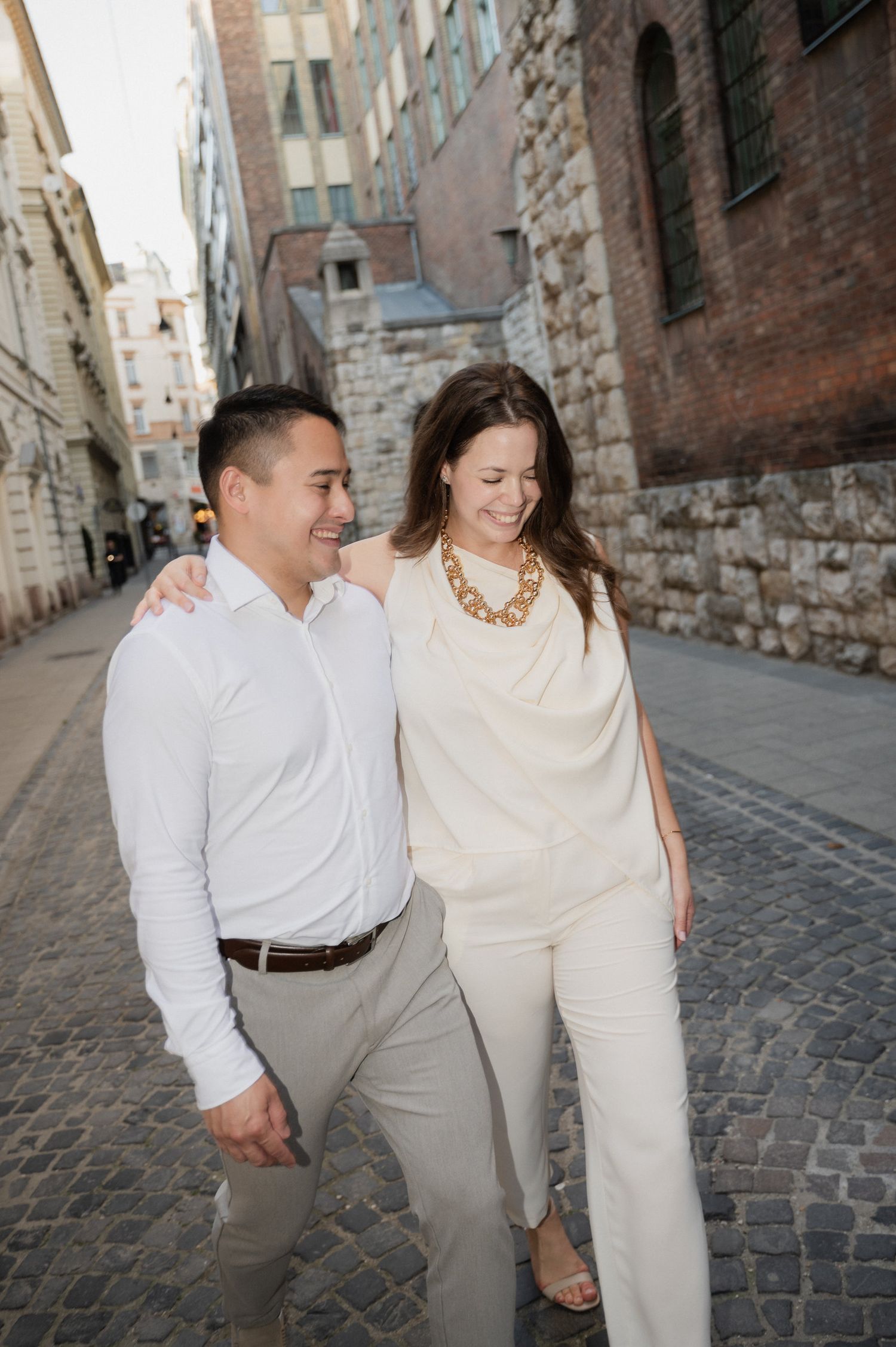 A couple in matching white outfits walking together through a charming European cobblestone street.