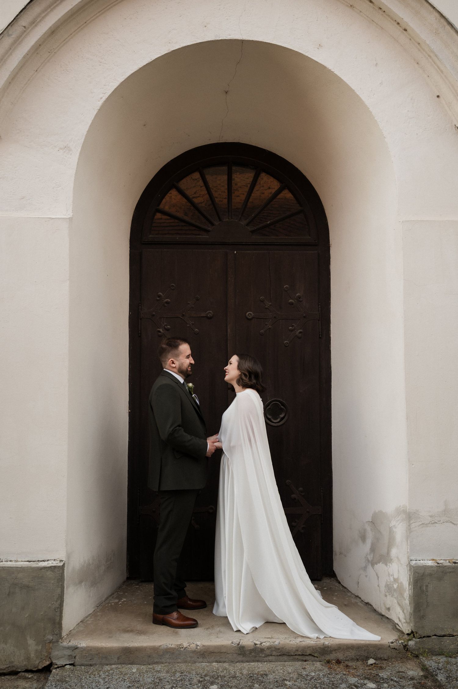 Wedding couple poses under white archway with dramatic dark wooden door backdrop.