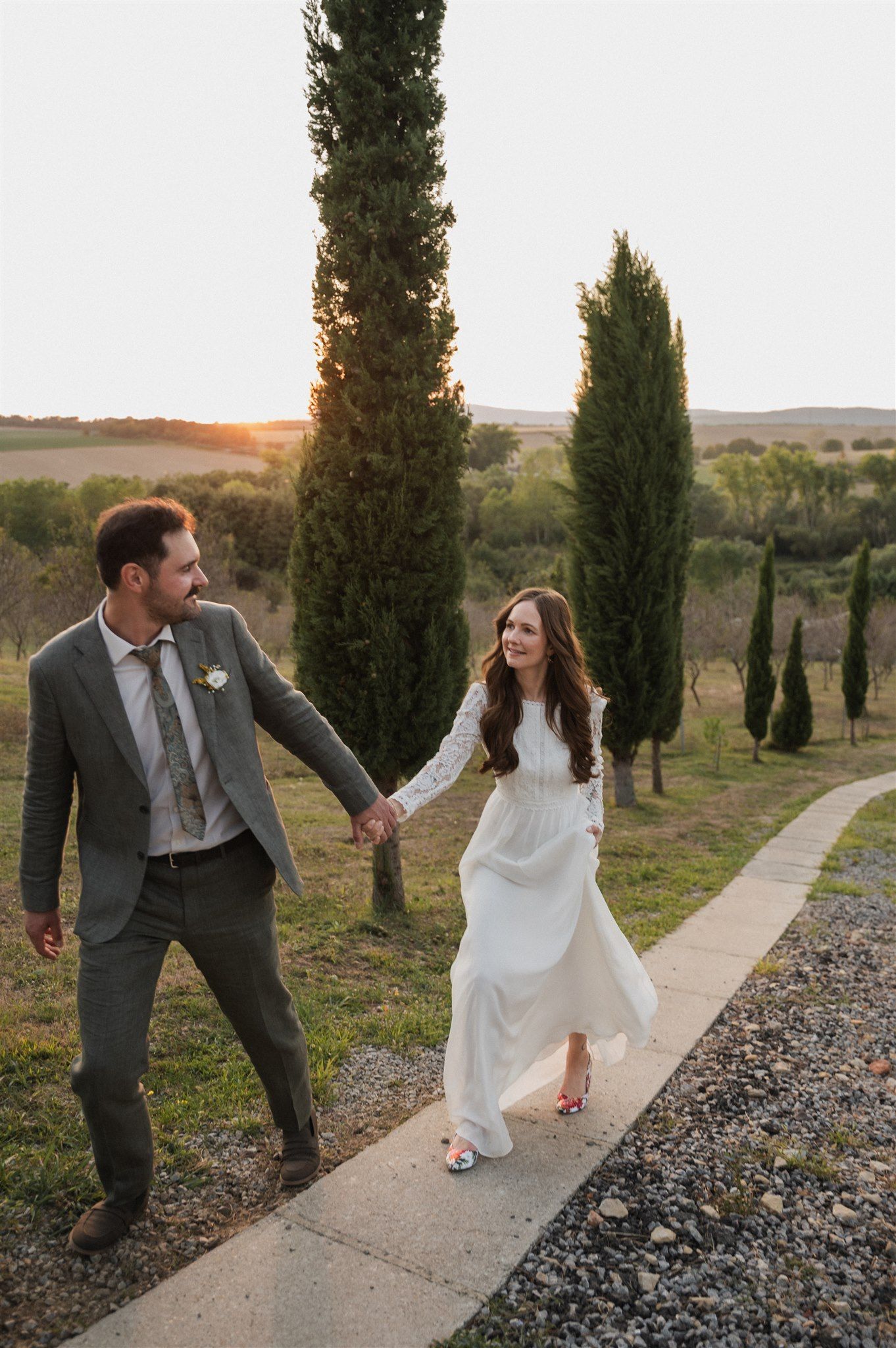 A couple walks along a path lined with cypress trees during sunset at a scenic countryside venue.