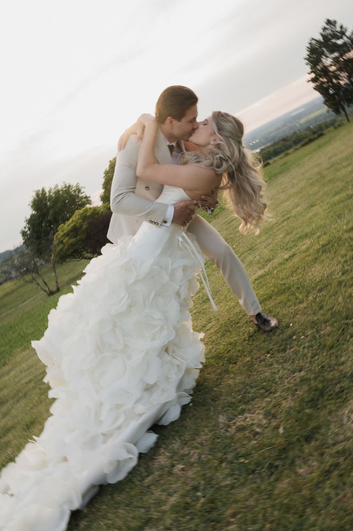 Groom dipping and kissing bride in a ruffled white wedding gown in an open green field at dusk.