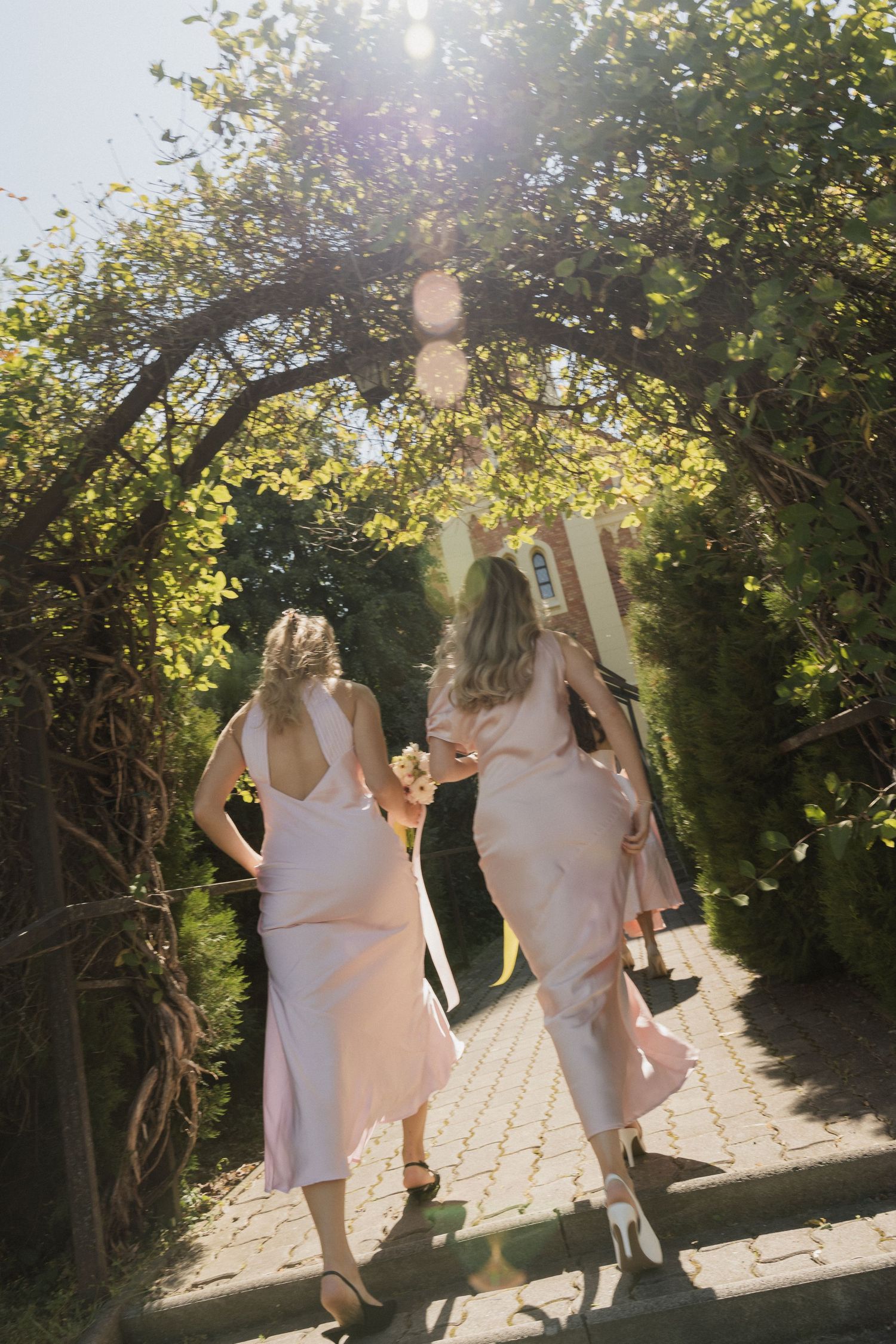 Two bridesmaids in pink dresses walk through a sunlit garden archway covered in lush green foliage.