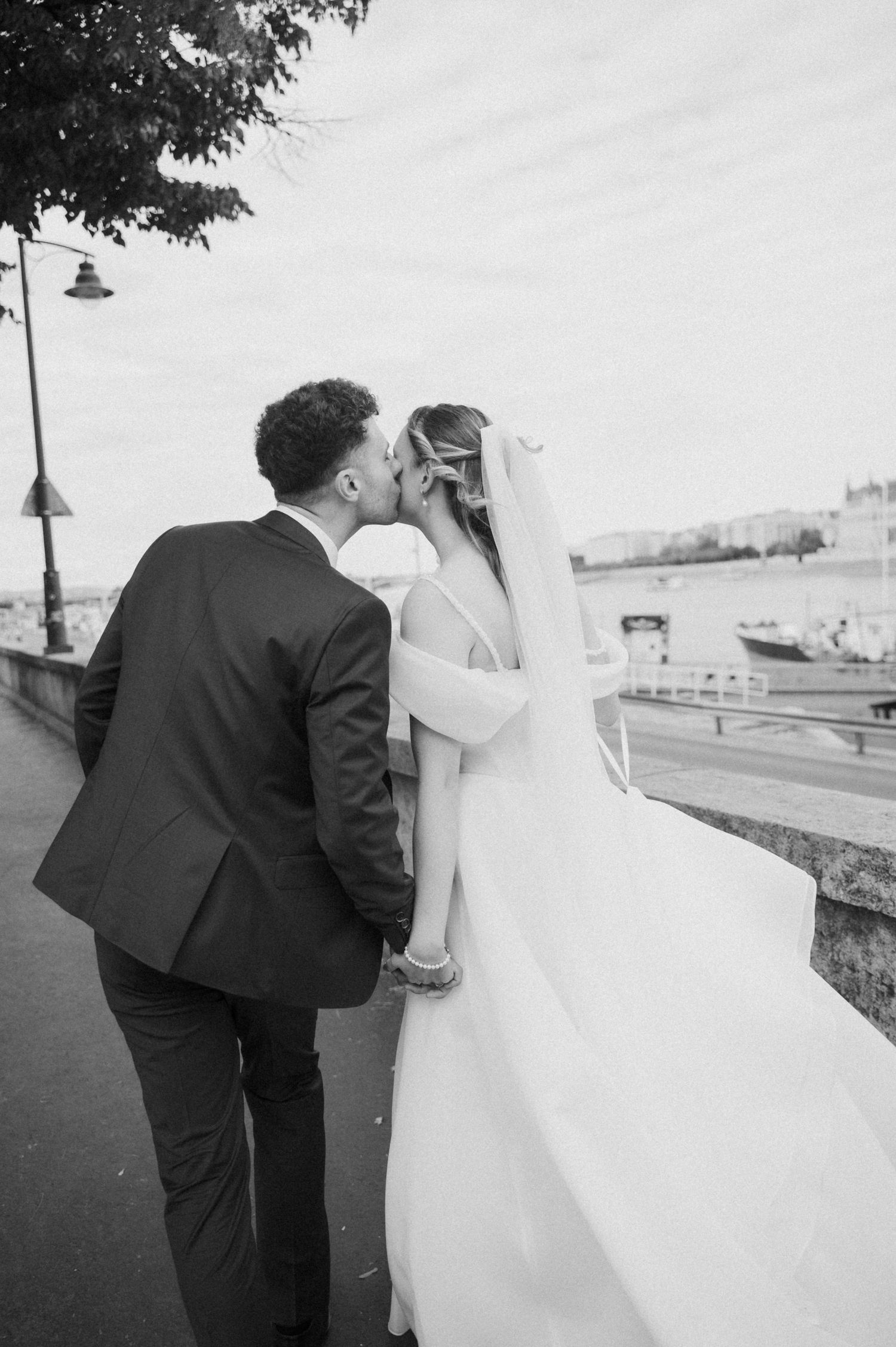 Bride and groom share romantic kiss on riverside promenade in black and white wedding photo.
