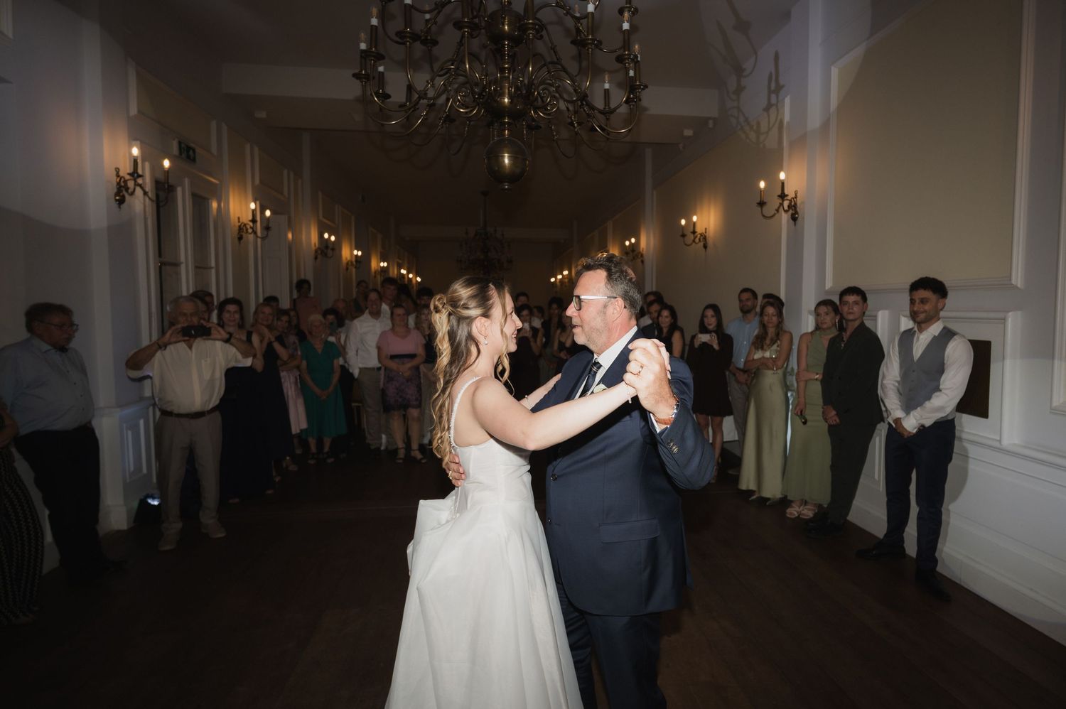 Bride and groom share first dance under chandelier while wedding guests watch in elegant ballroom.