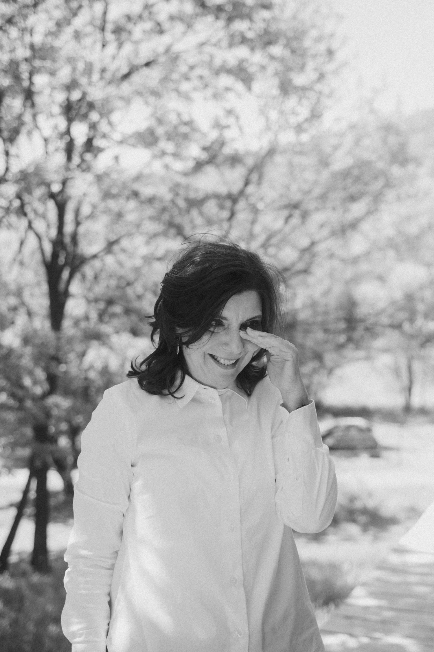 A figure in a white shirt stands outdoors against a soft blurred background of trees and sunlight.