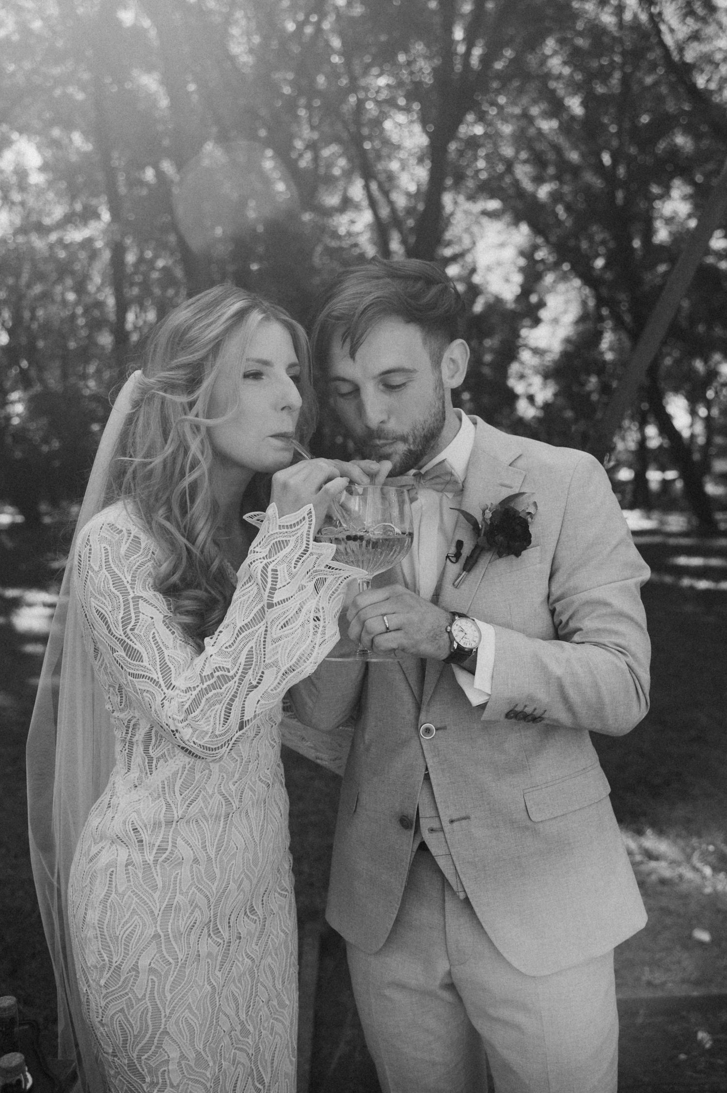A couple in wedding attire sharing a drink together outdoors in a black and white photo.