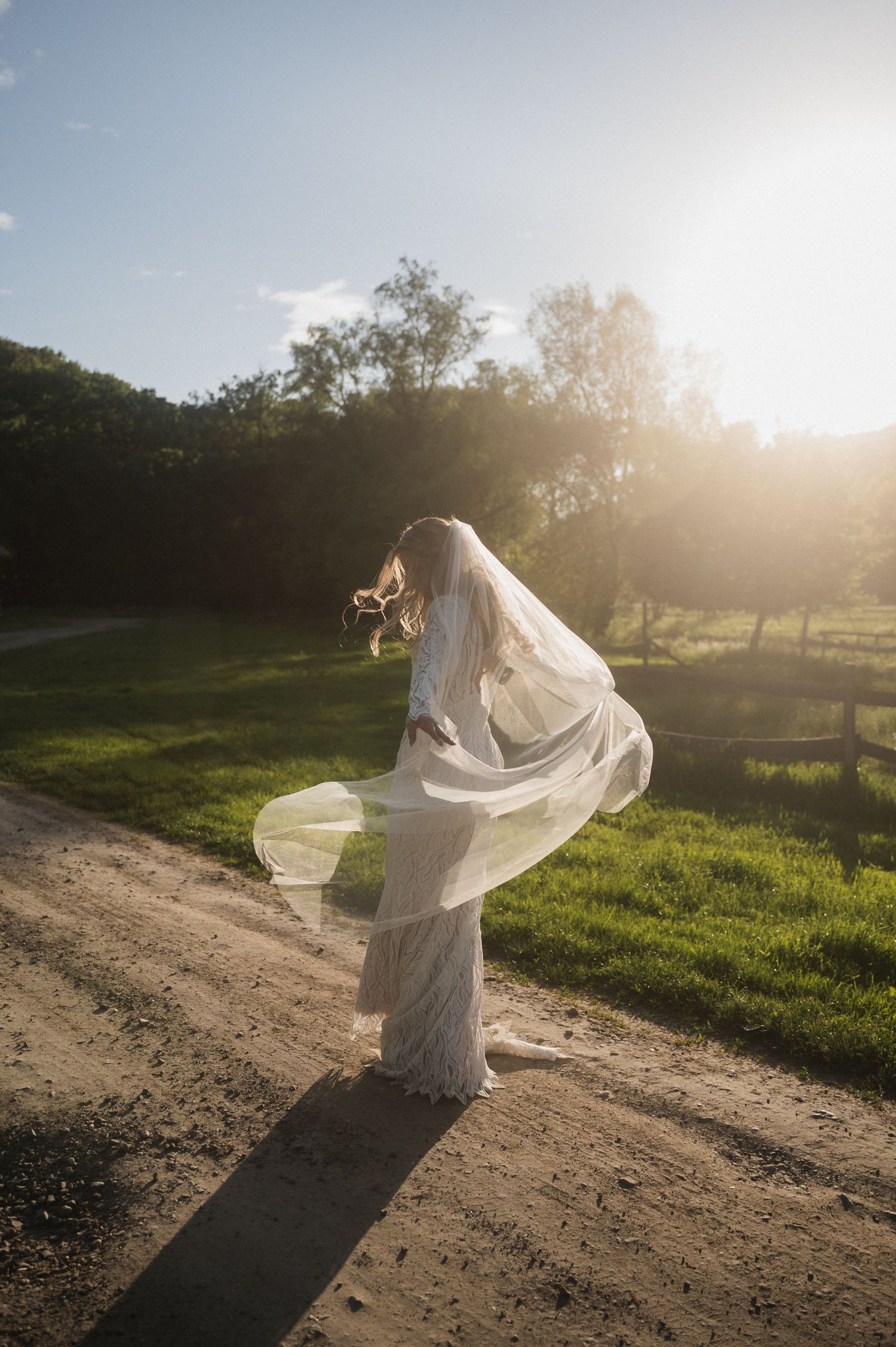 A flowing white wedding dress billows in the breeze as sunlight streams through trees on a dirt path at sunset.