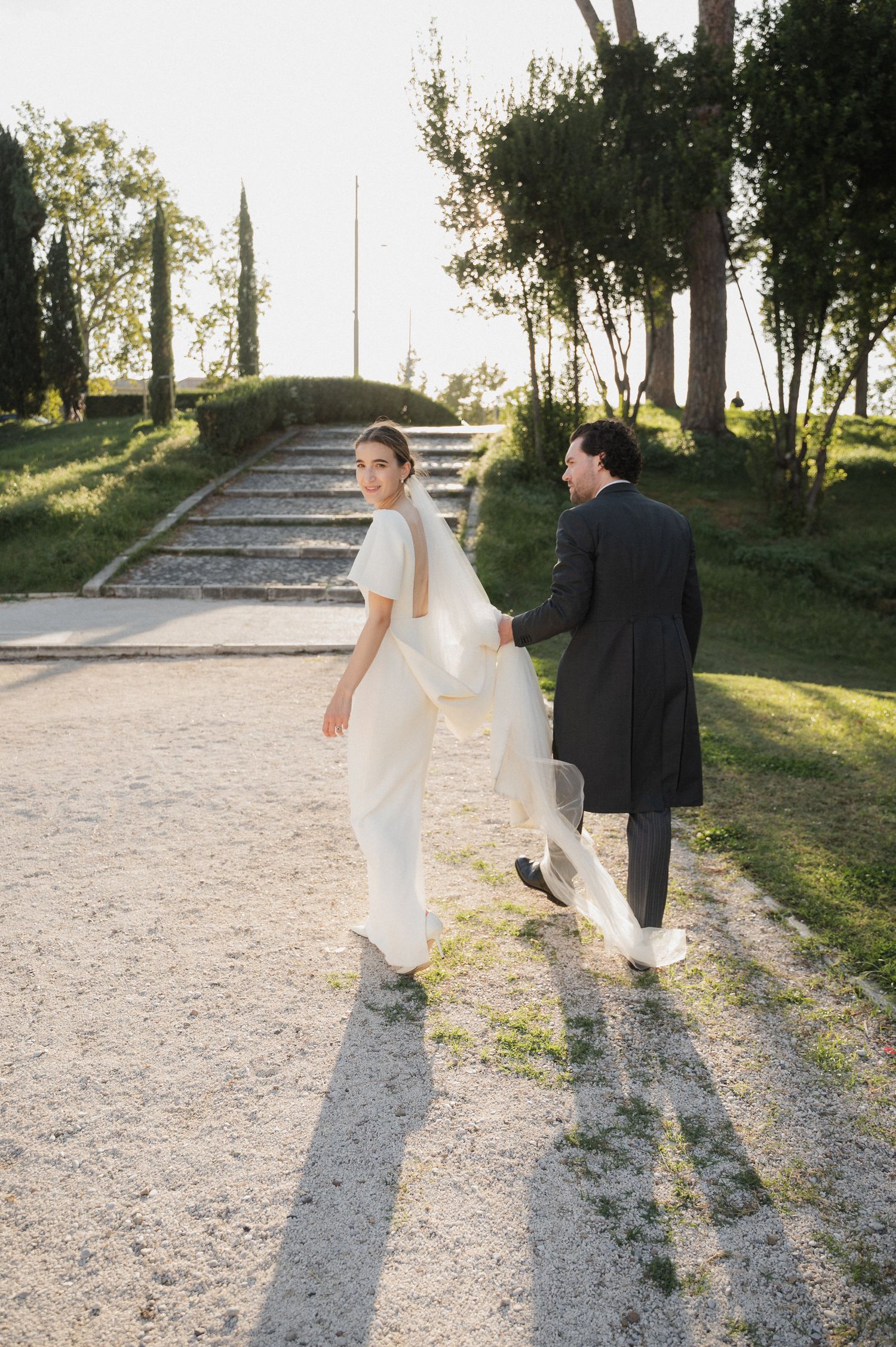 Newlyweds walk together casting long shadows on a sunlit gravel path.