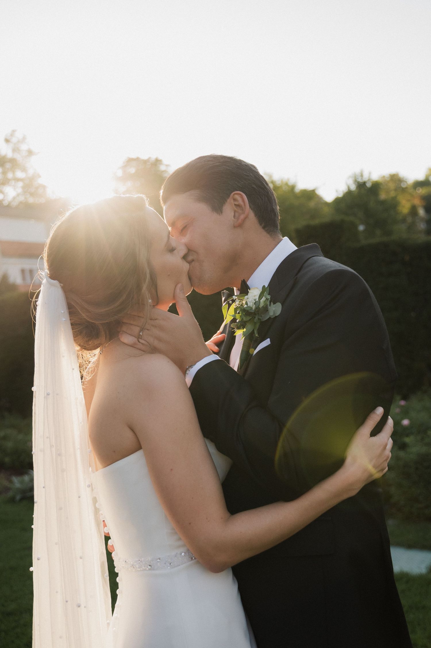 A romantic wedding couple embraces in golden sunlight during sunset while sharing an intimate kiss.