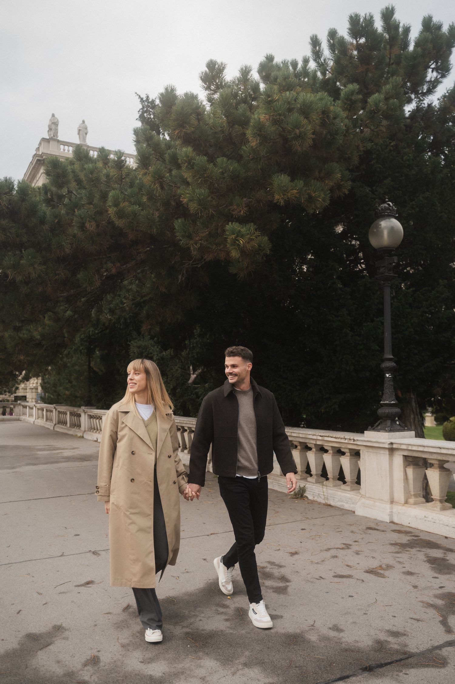 A couple walking together on a stone bridge path wearing beige coats and dark clothing on an overcast day.