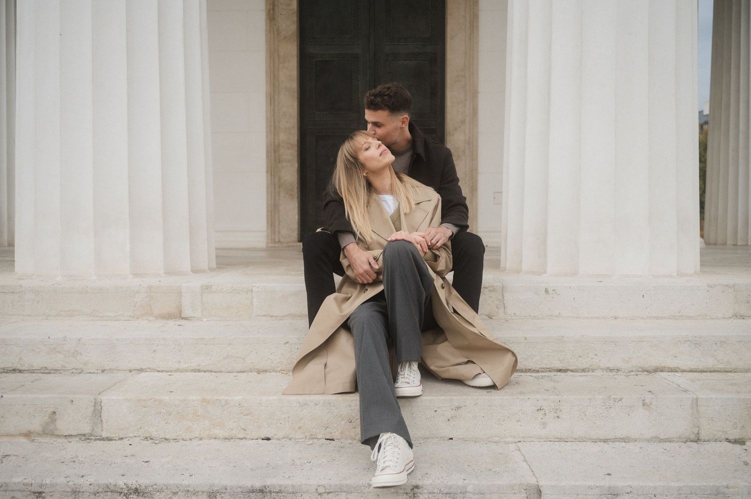 A couple poses playfully in front of white marble columns in a series of romantic engagement photos.