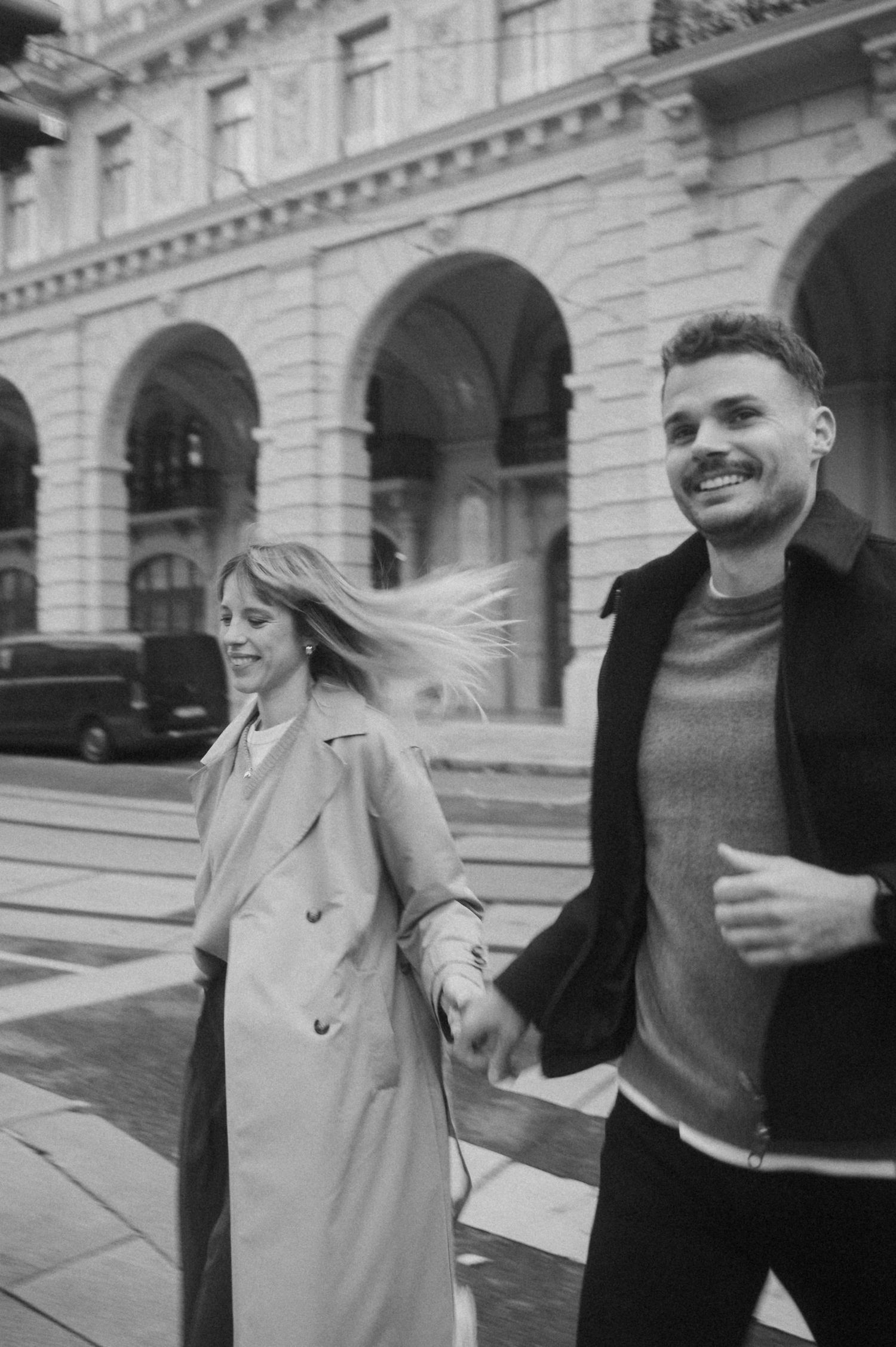 A couple walks together in front of a grand historic building with arched architecture in black and white.