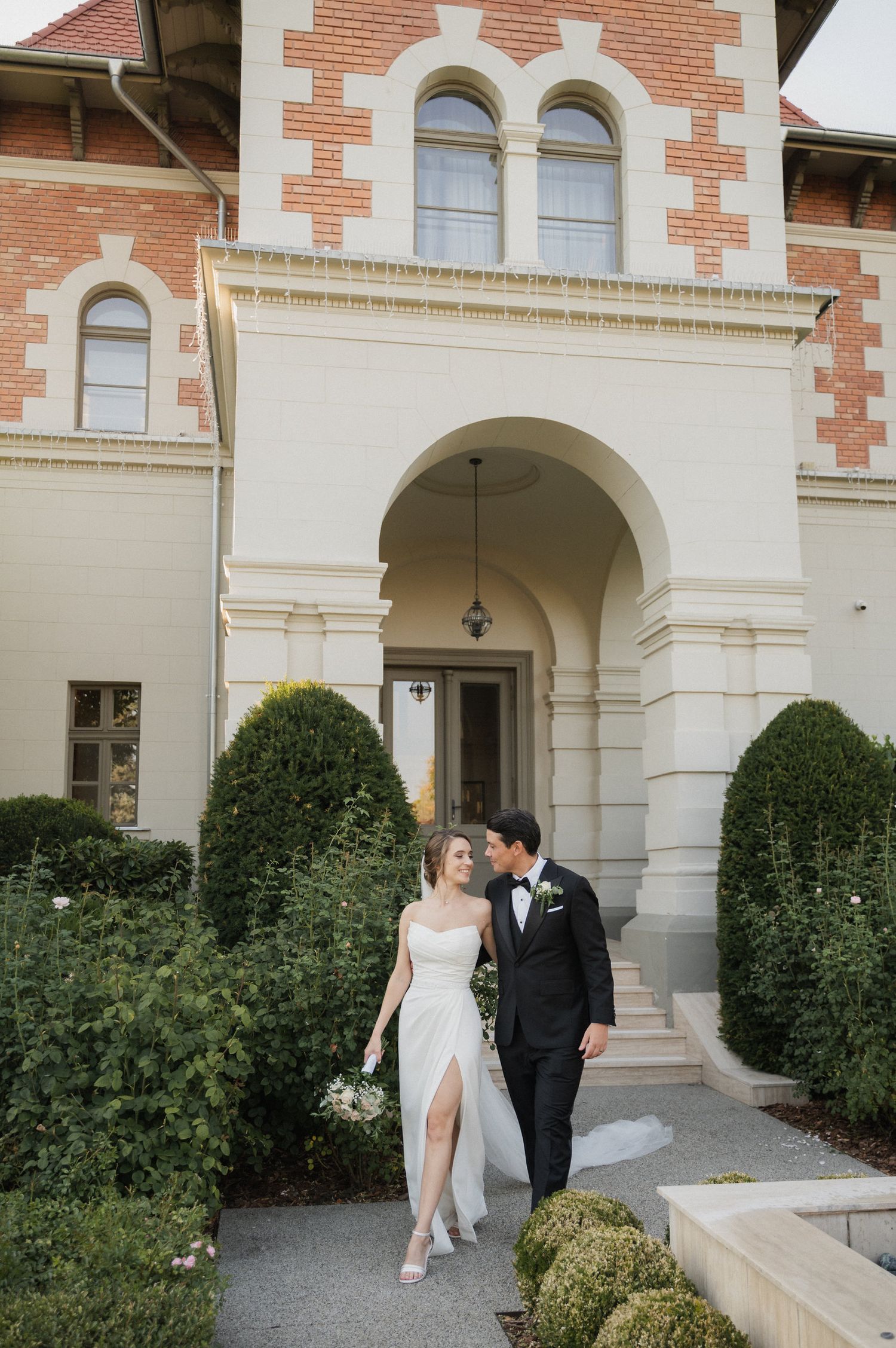 A couple in wedding attire walking together through an elegant archway of a brick and stone building.