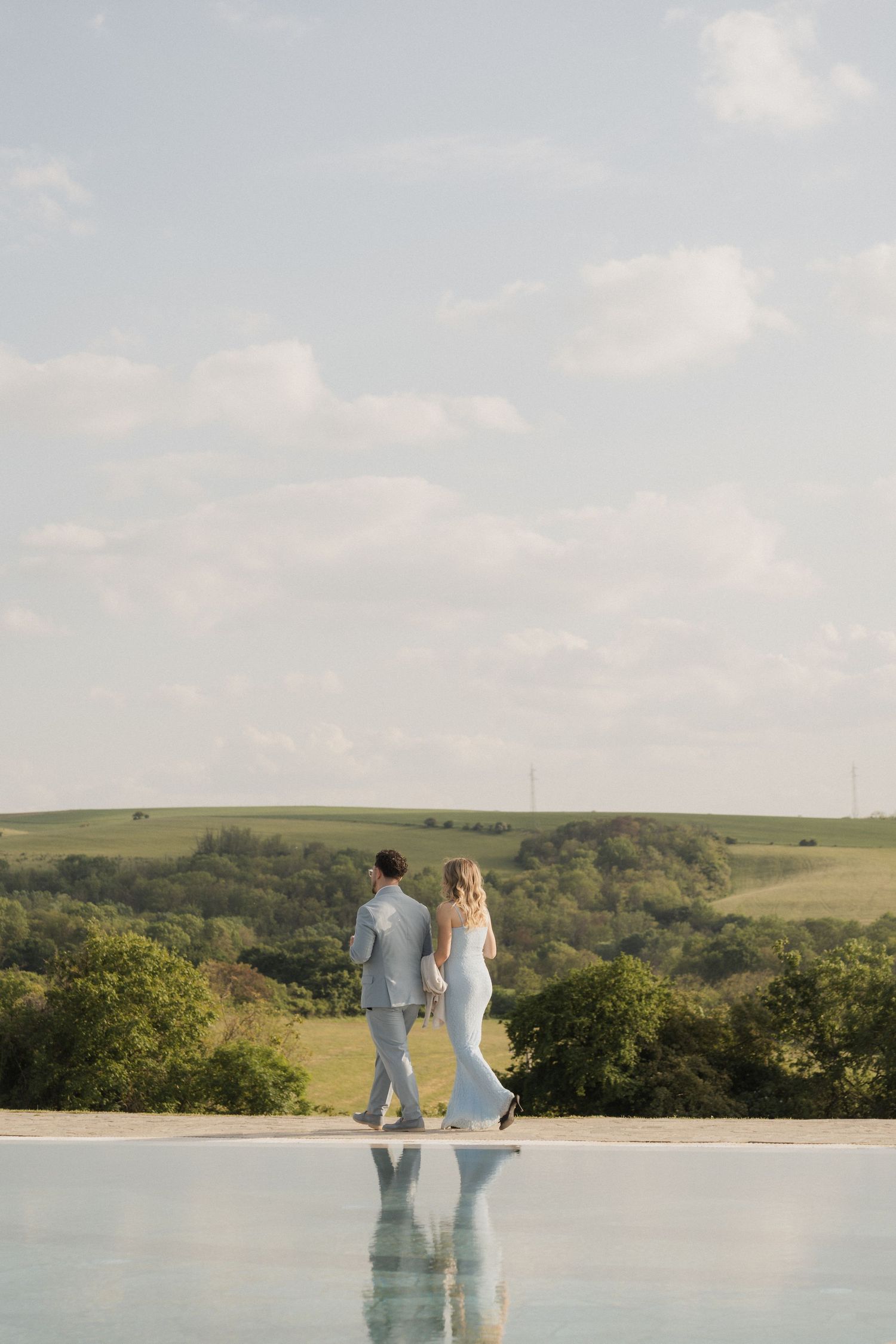 Bride and groom holding hands by an infinity pool overlooking a lush green countryside landscape.