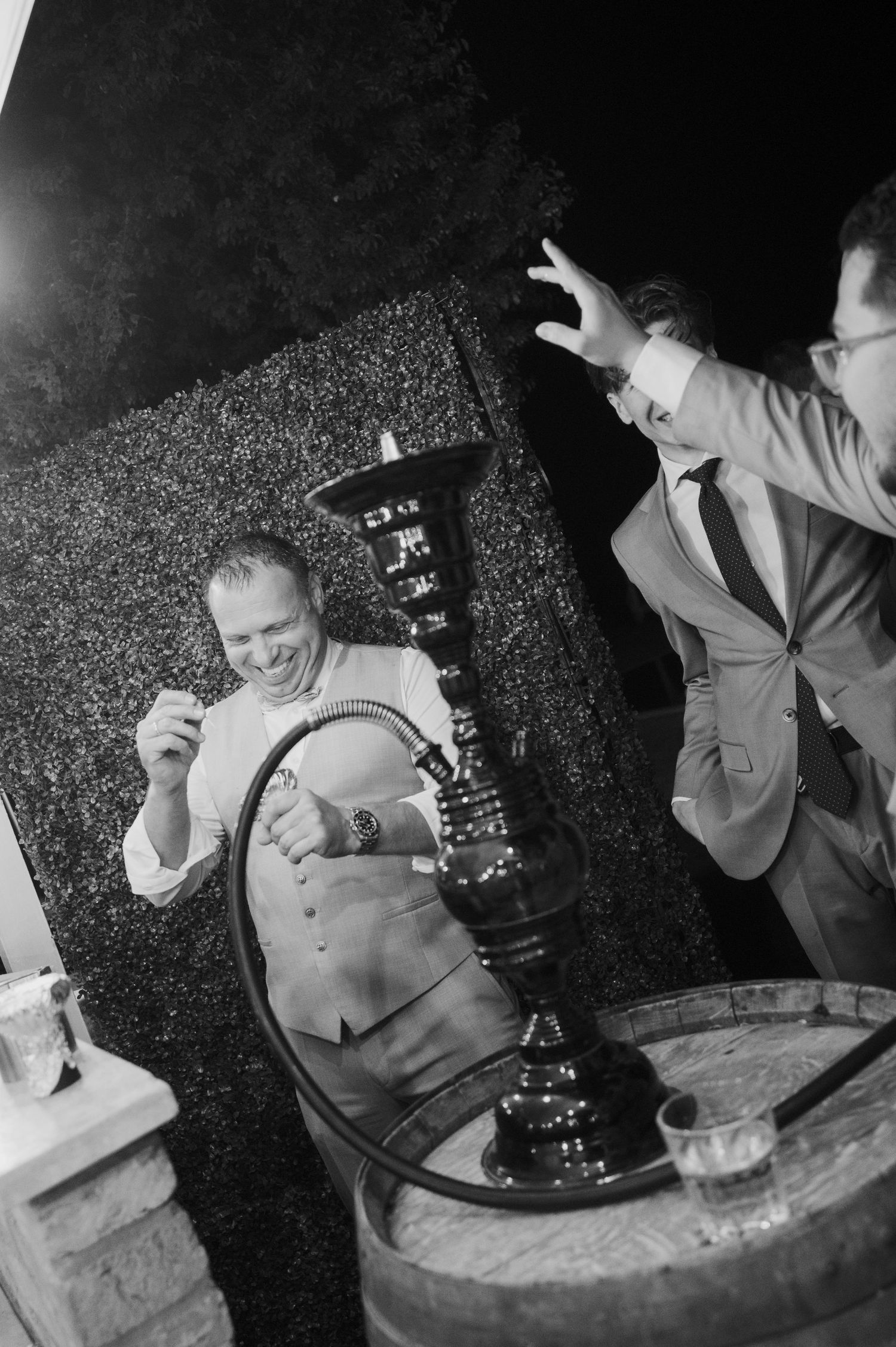 Two men laugh beside a large hookah pipe at an outdoor nighttime event in black and white.