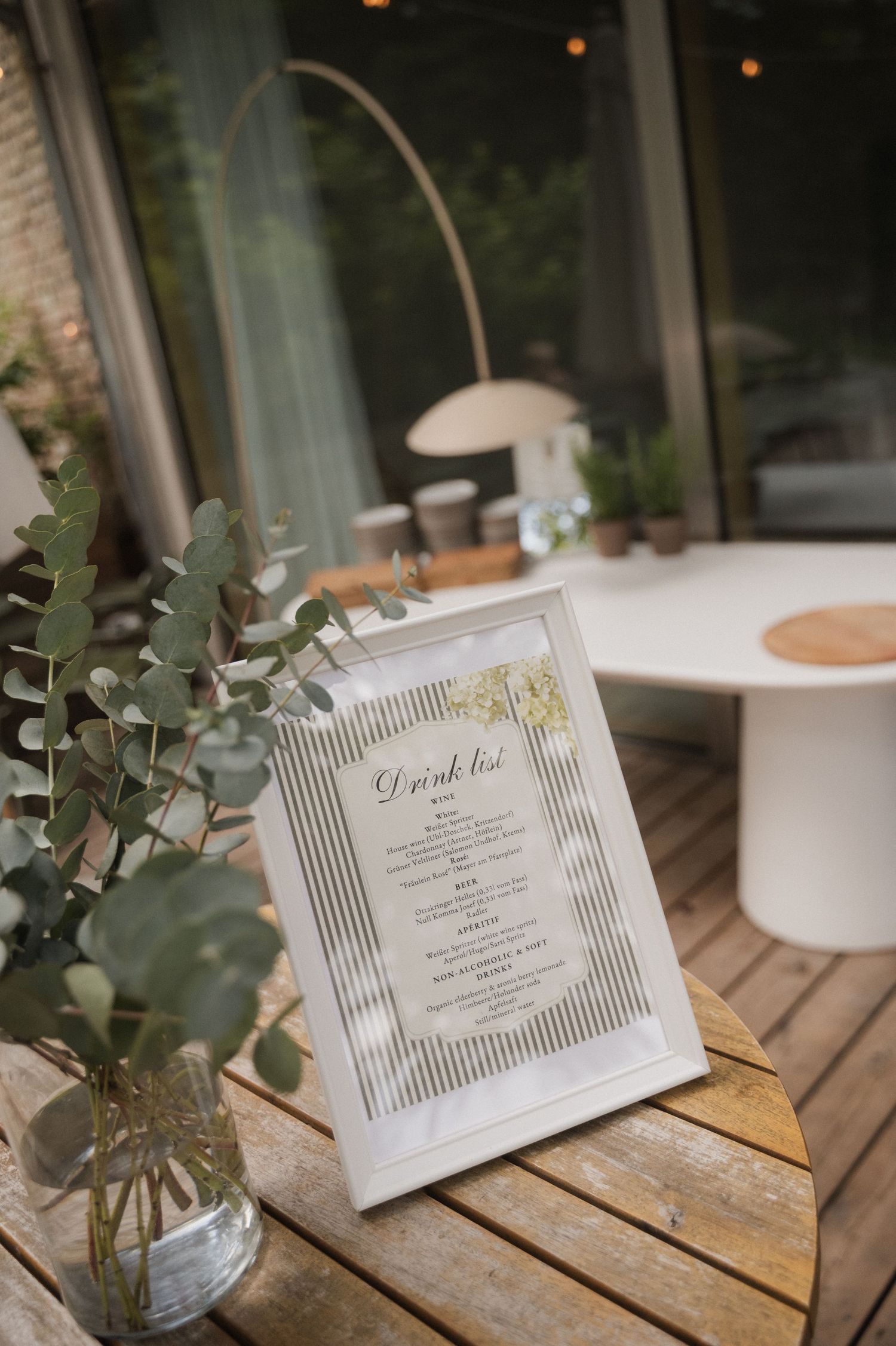 Elegant framed drink list menu displayed on wooden table with eucalyptus greenery at an outdoor wedding reception.