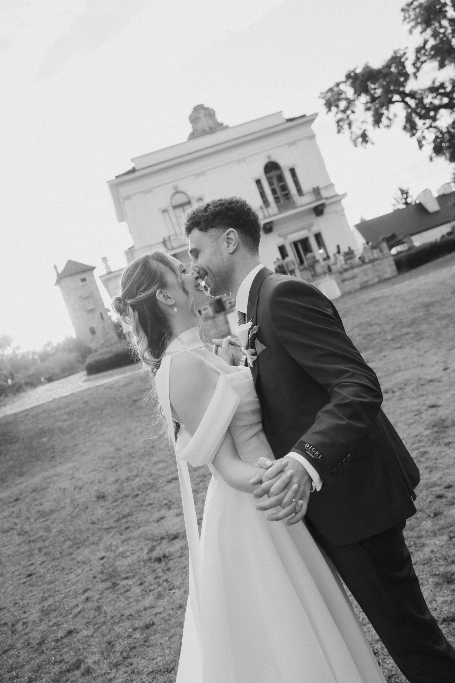Black and white photo of a bride and groom sharing a romantic moment, holding hands in front of an elegant venue.