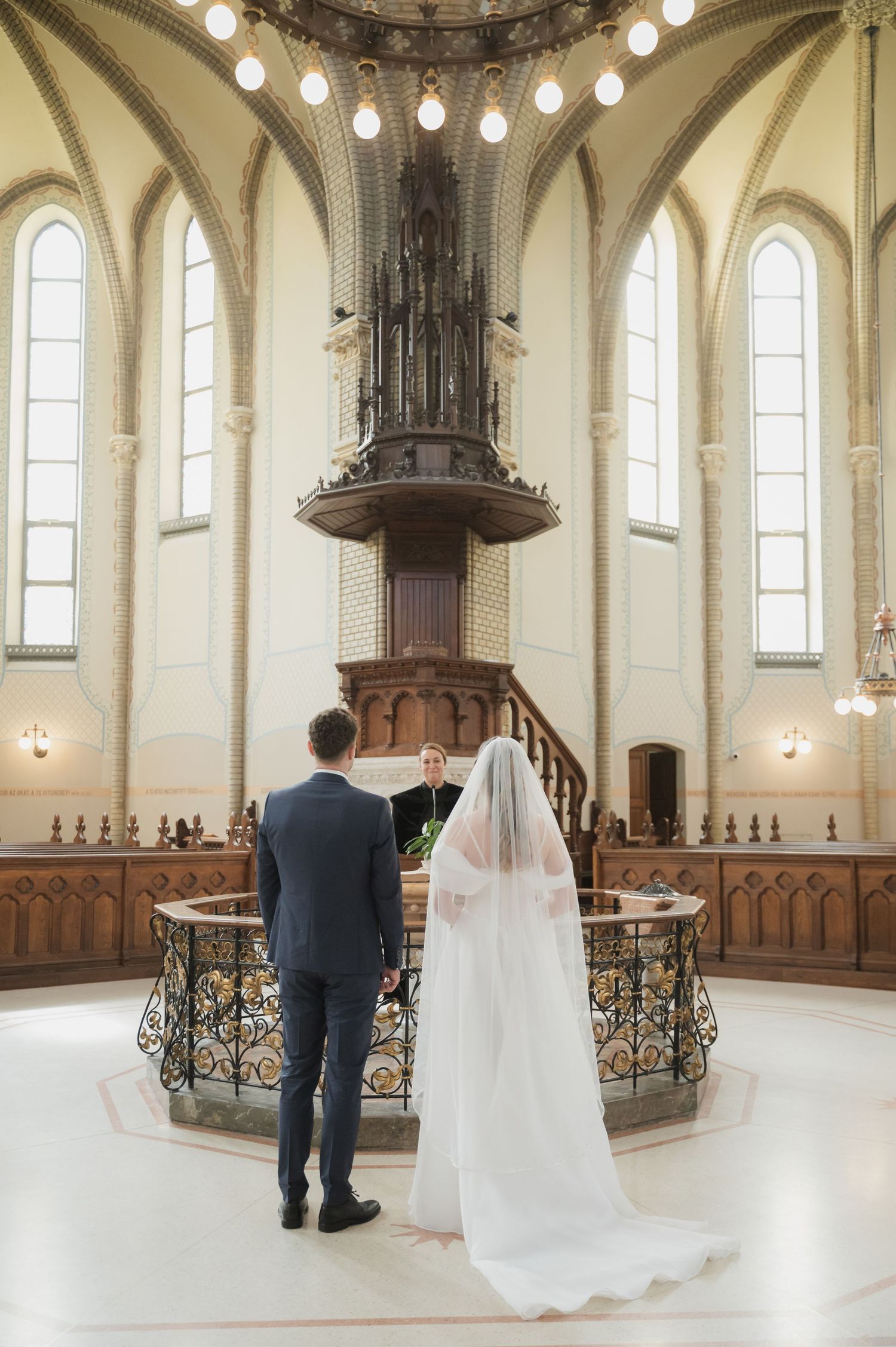 Bride and groom stand before officiant at elegant church altar with ornate wooden pulpit during wedding ceremony.