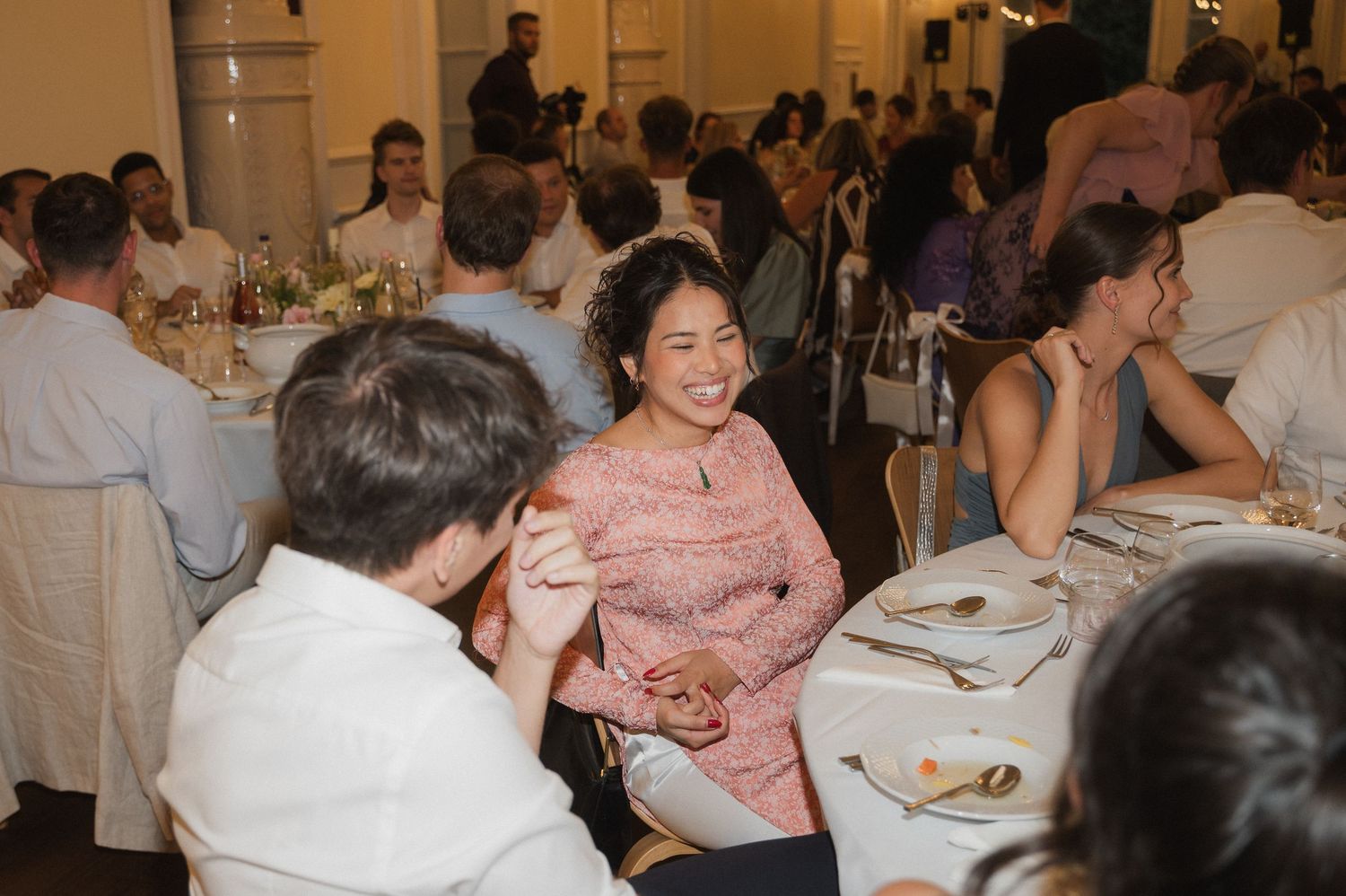 Wedding reception guests laughing and conversing at elegantly set dinner tables indoors.
