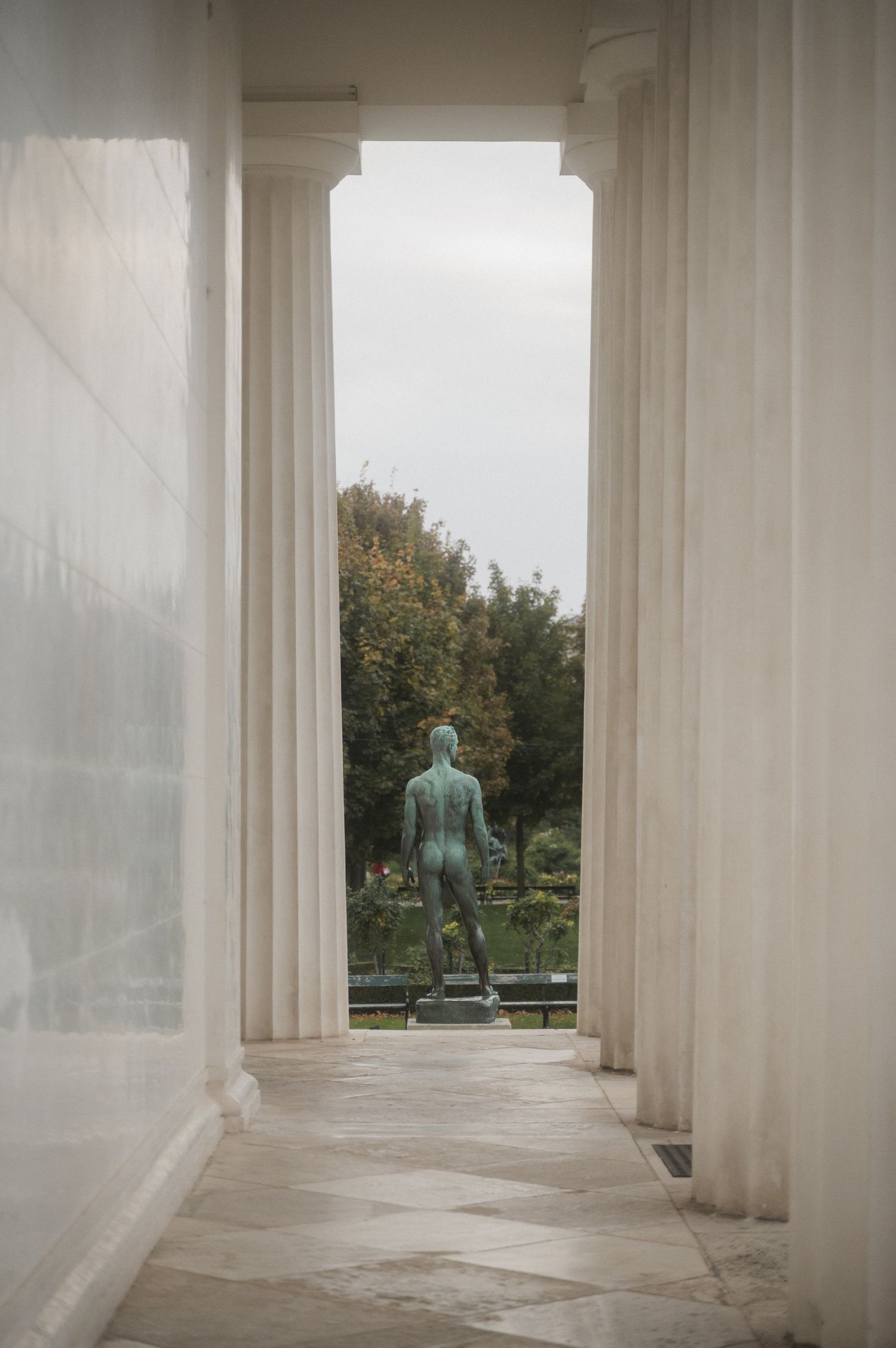 White marble columns frame a distant view of trees and a statue in a serene garden setting.