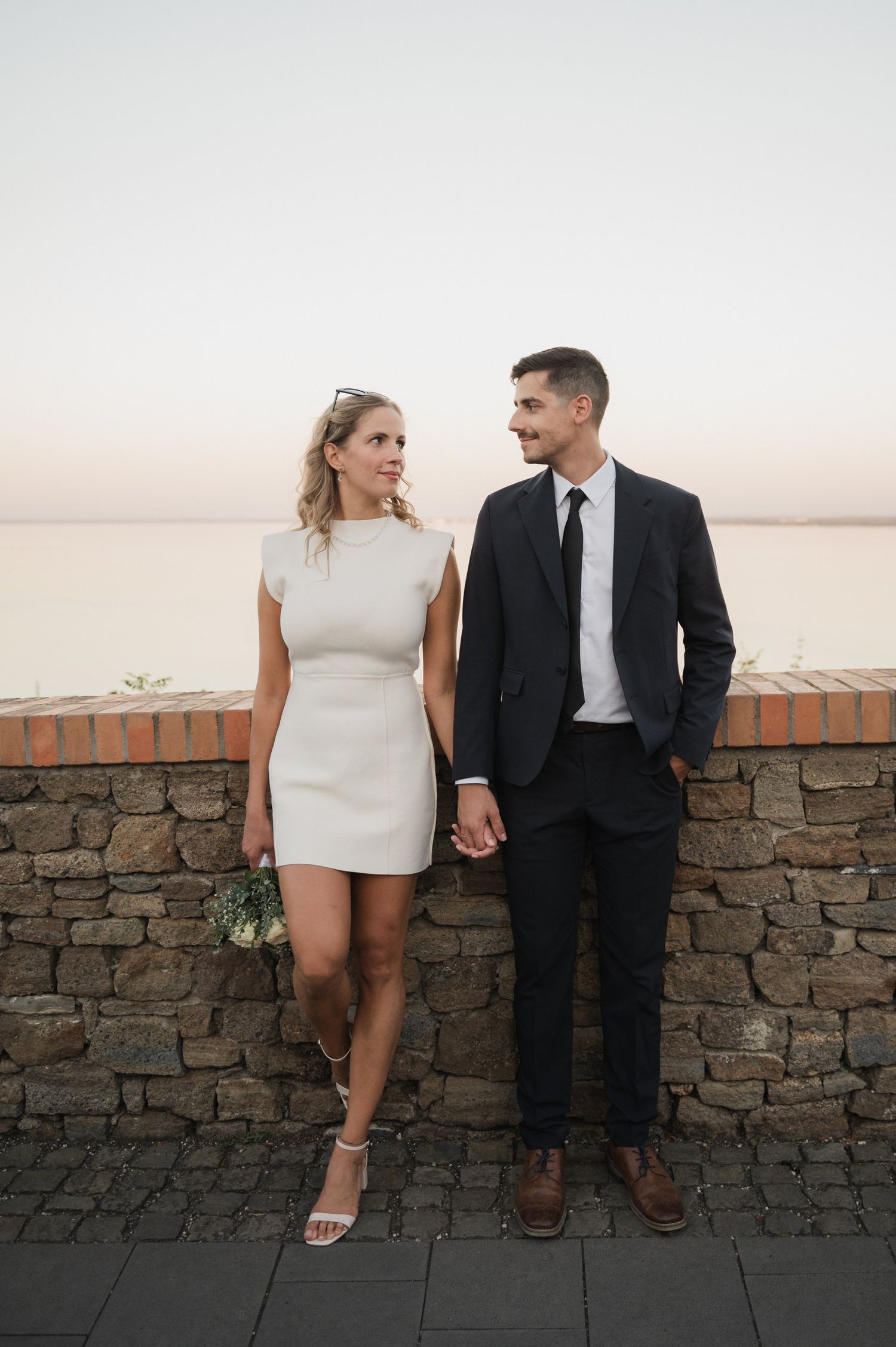 A couple in contrasting black and white outfits sit together on a stone wall at sunset.