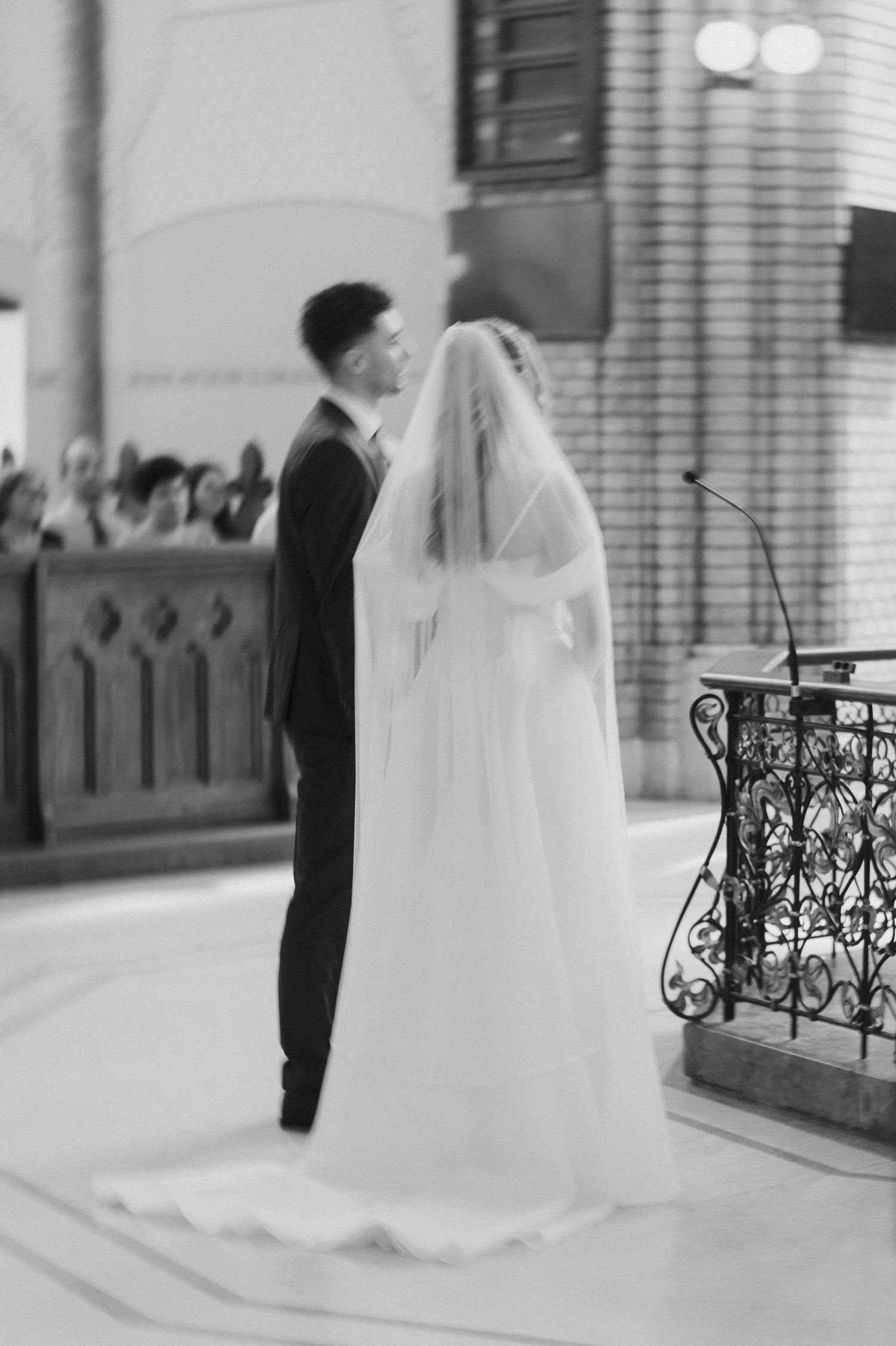 Black and white photo of bride in flowing veil and groom standing together at church altar during wedding ceremony.