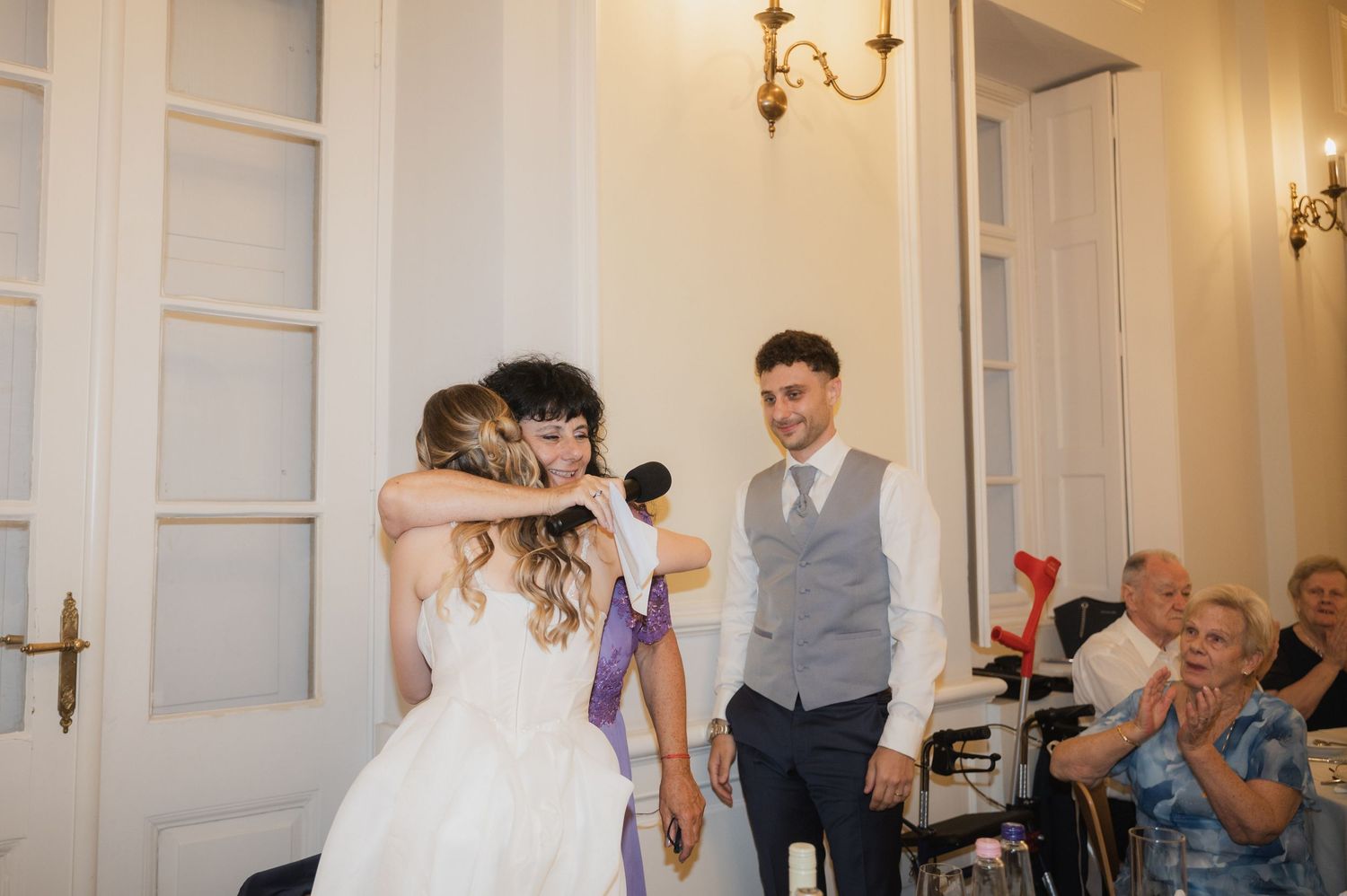 Bride hugs guest while groom watches and wedding attendees applaud during joyful reception celebration.
