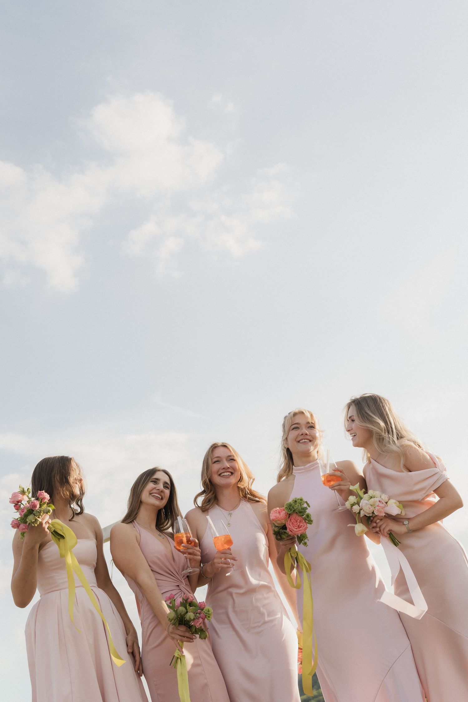 Five bridesmaids in pink dresses laugh and celebrate outdoors holding bouquets and champagne glasses.