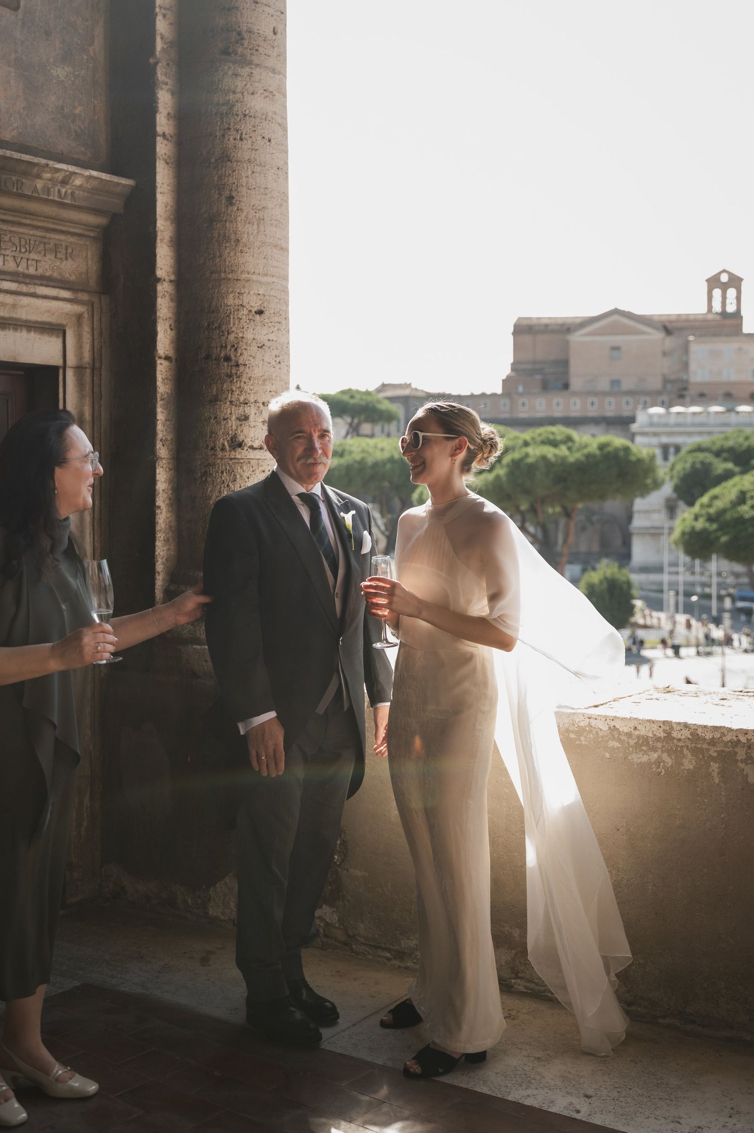 Elegant wedding party gathering under historic stone columns with sunlit views of Rome.