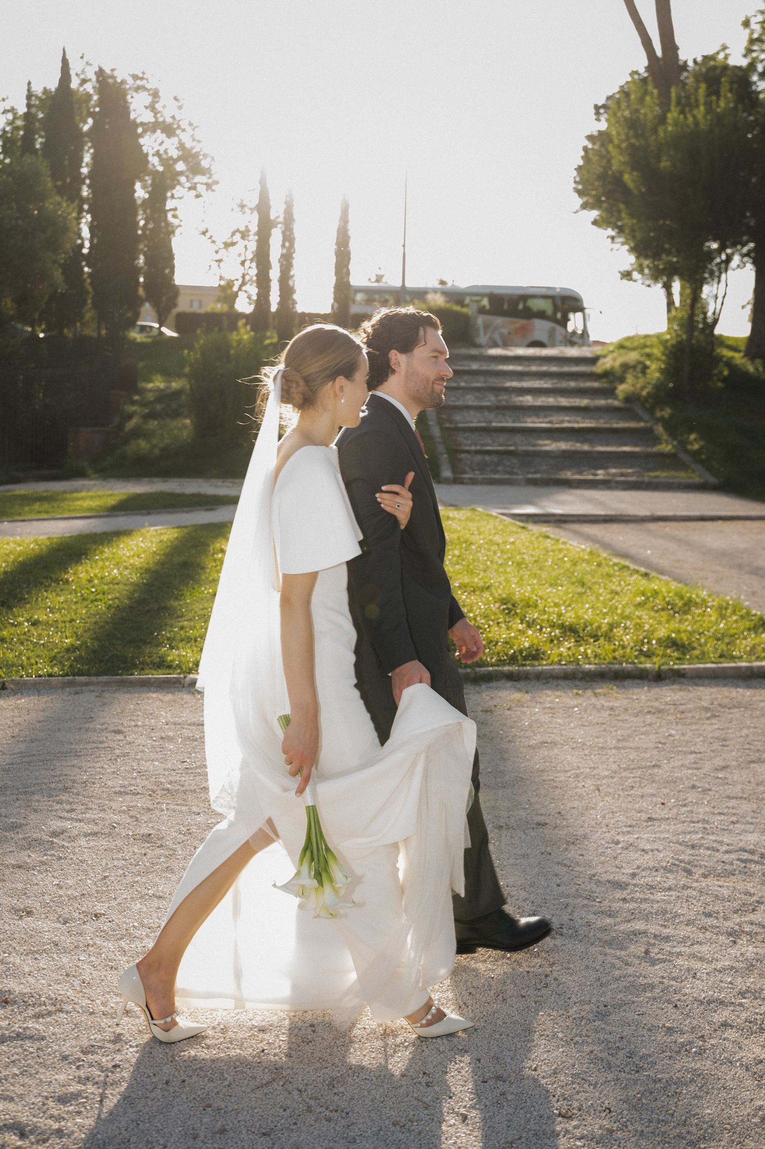 A couple in wedding attire walks together on a sunlit path casting long shadows.