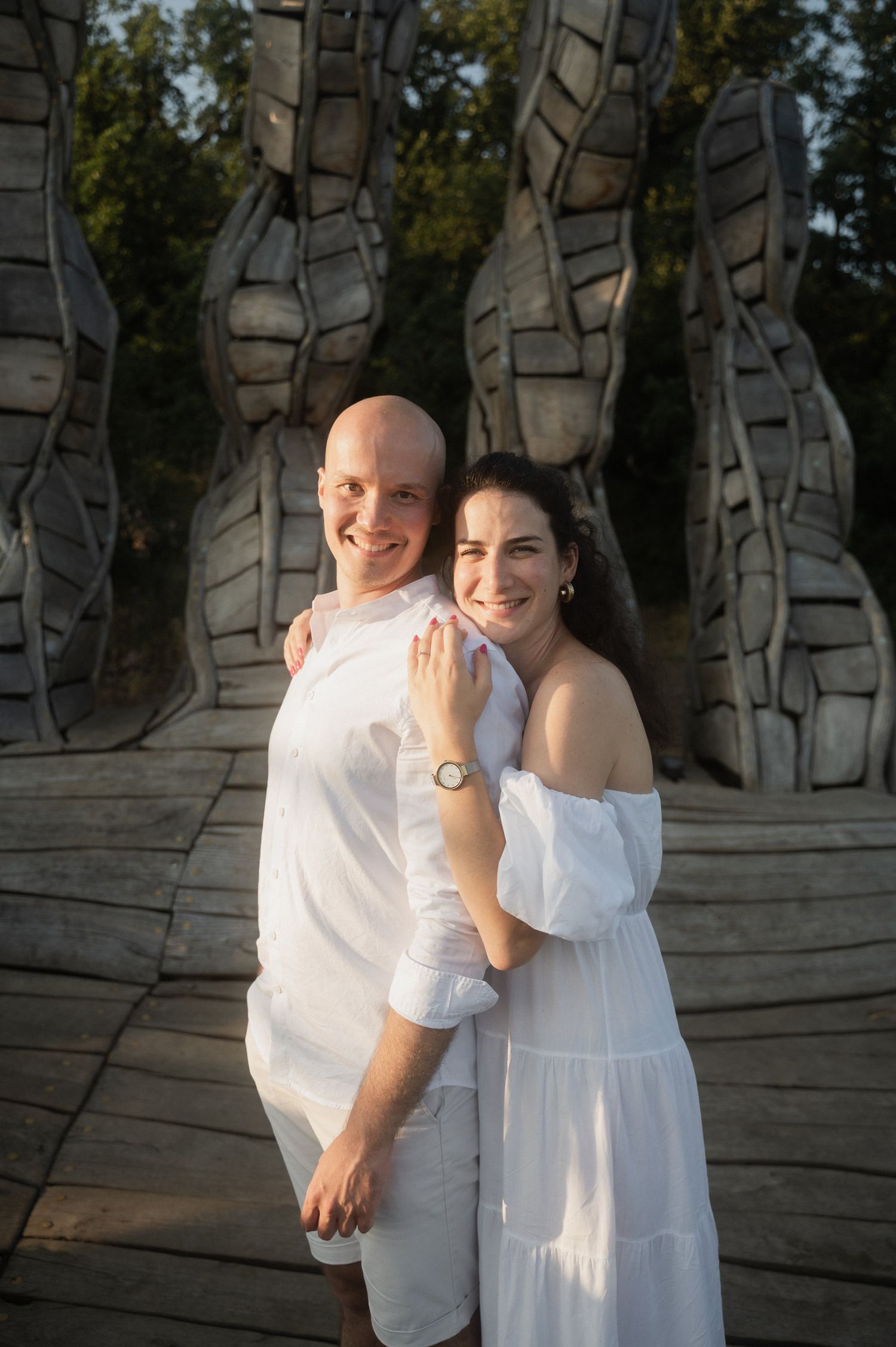 A couple in white outfits poses together in front of a modern art installation with tall sculptural forms.