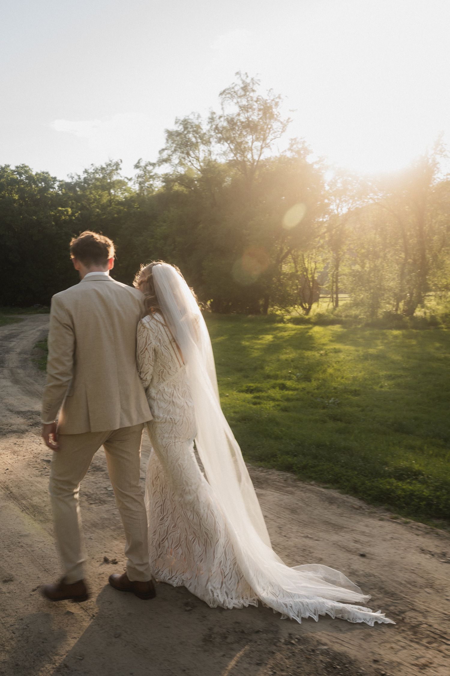 A couple in wedding attire walks down a sunlit dirt path through a grassy field during golden hour.