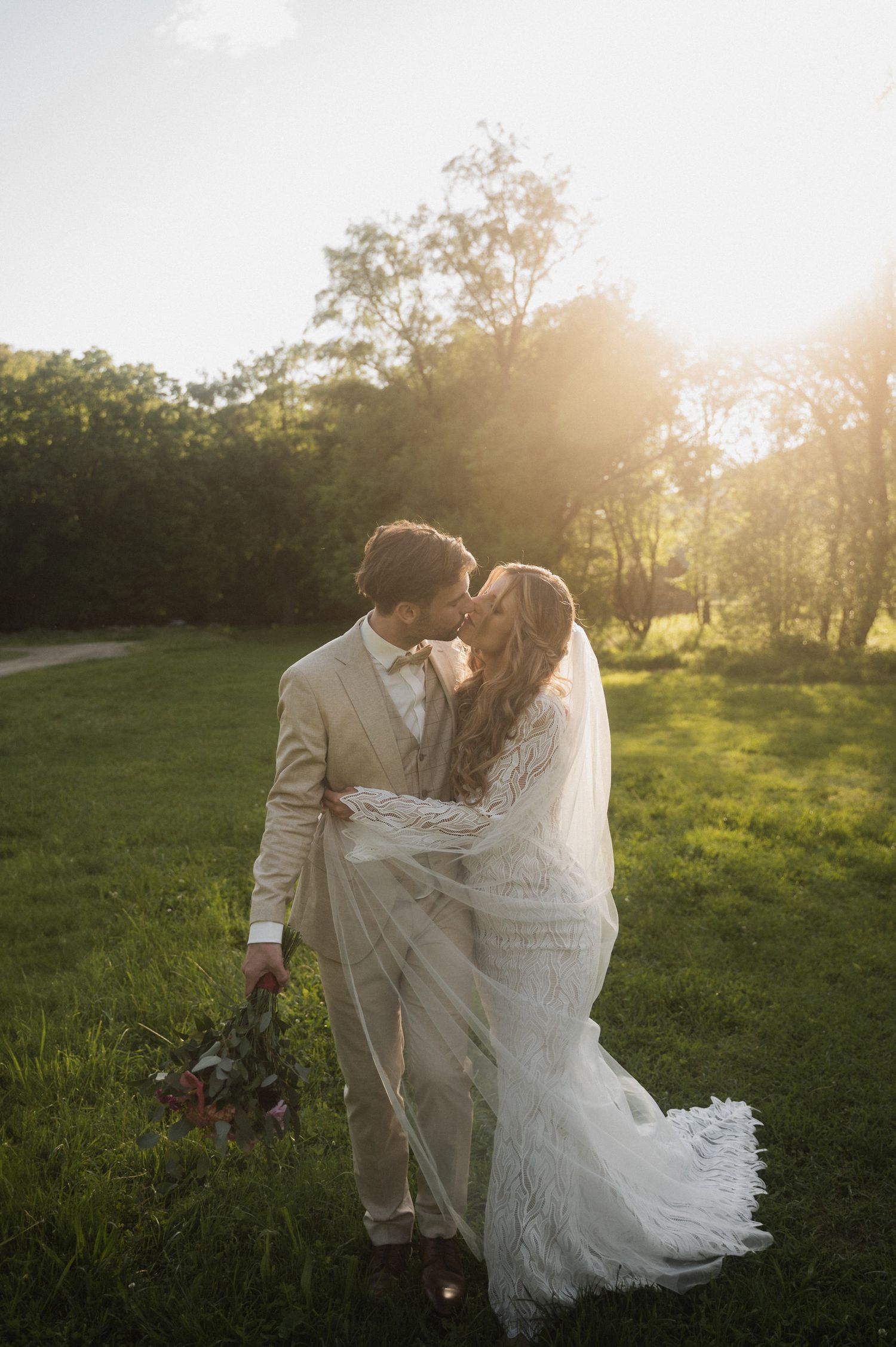 A romantic wedding couple embraces in a sun-drenched meadow at golden hour with trees in the background.