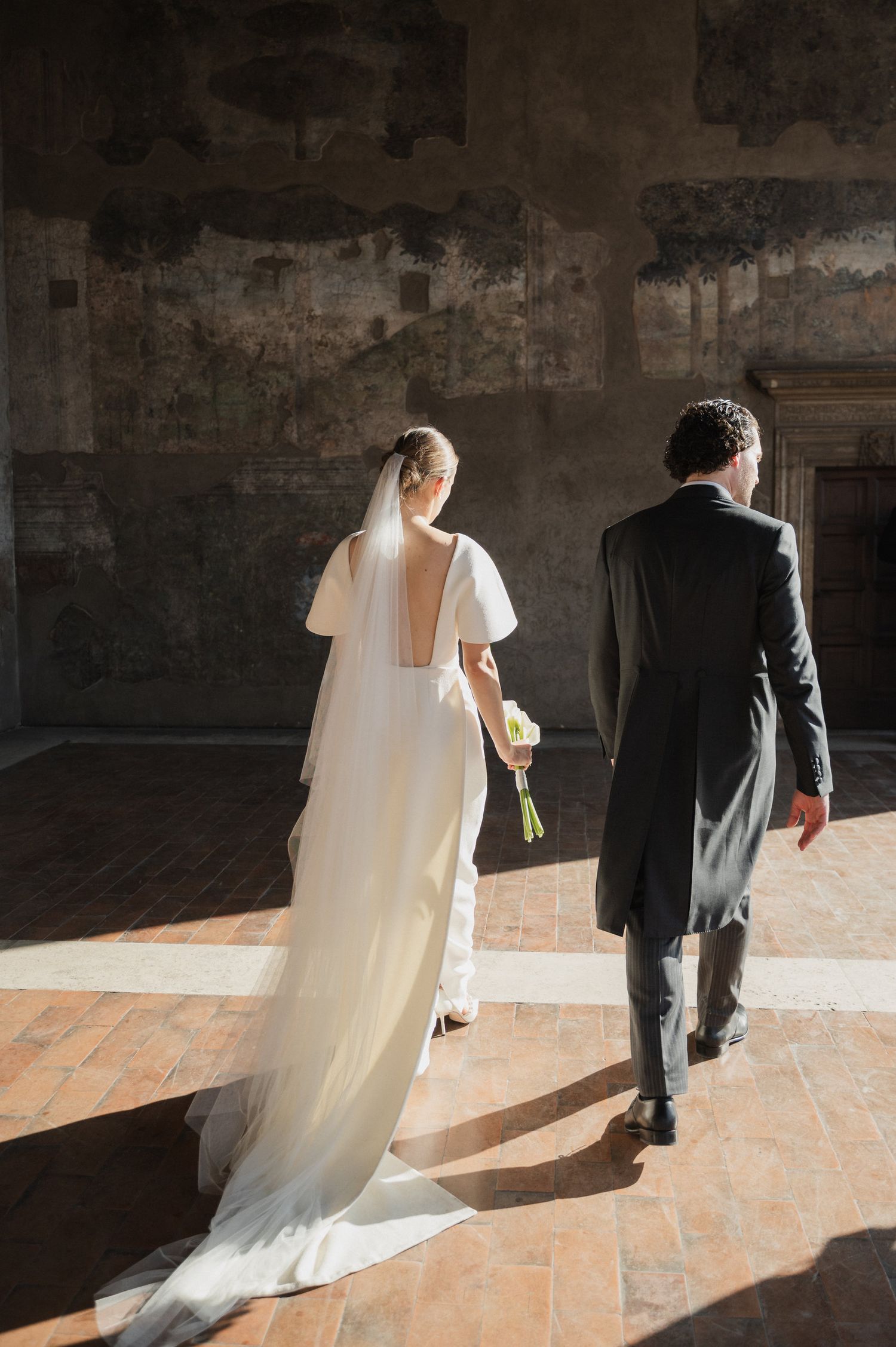 Silhouetted couple walking through sunlit stone corridor in wedding attire.
