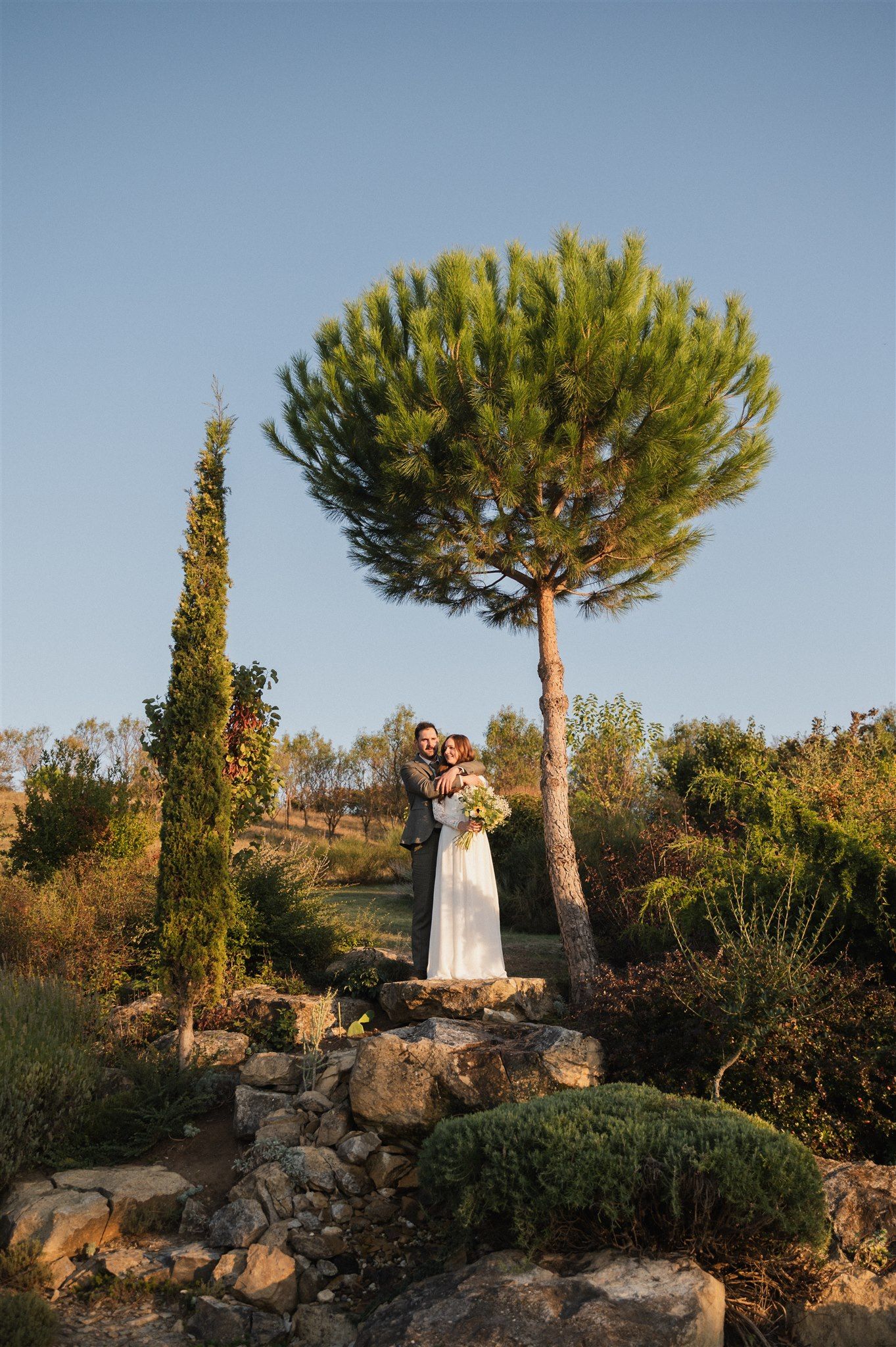 A romantic wedding couple stands on stone steps beneath a tall Mediterranean pine tree at sunset.