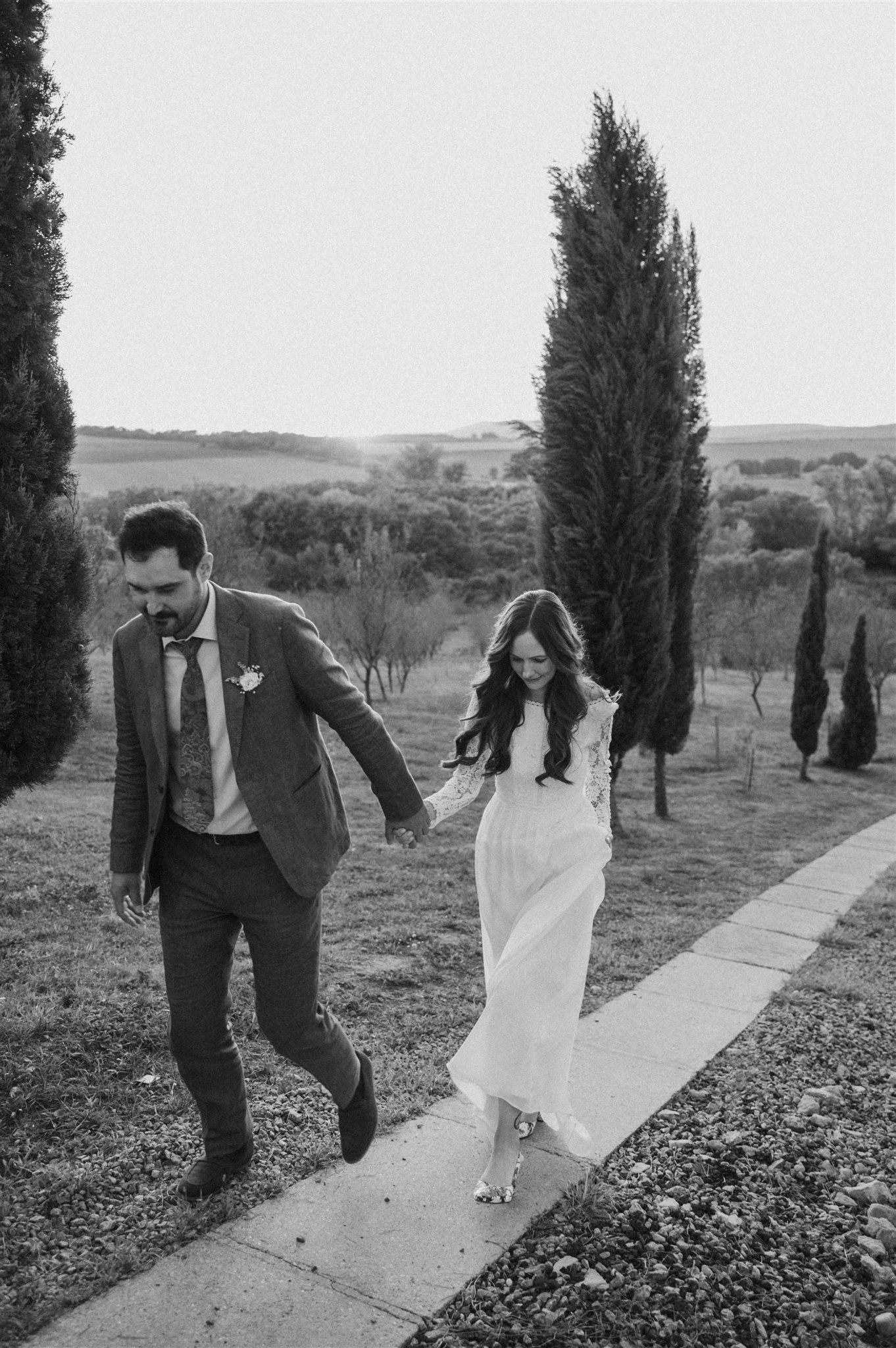 A couple joyfully walks along a pathway surrounded by tall cypress trees in black and white.