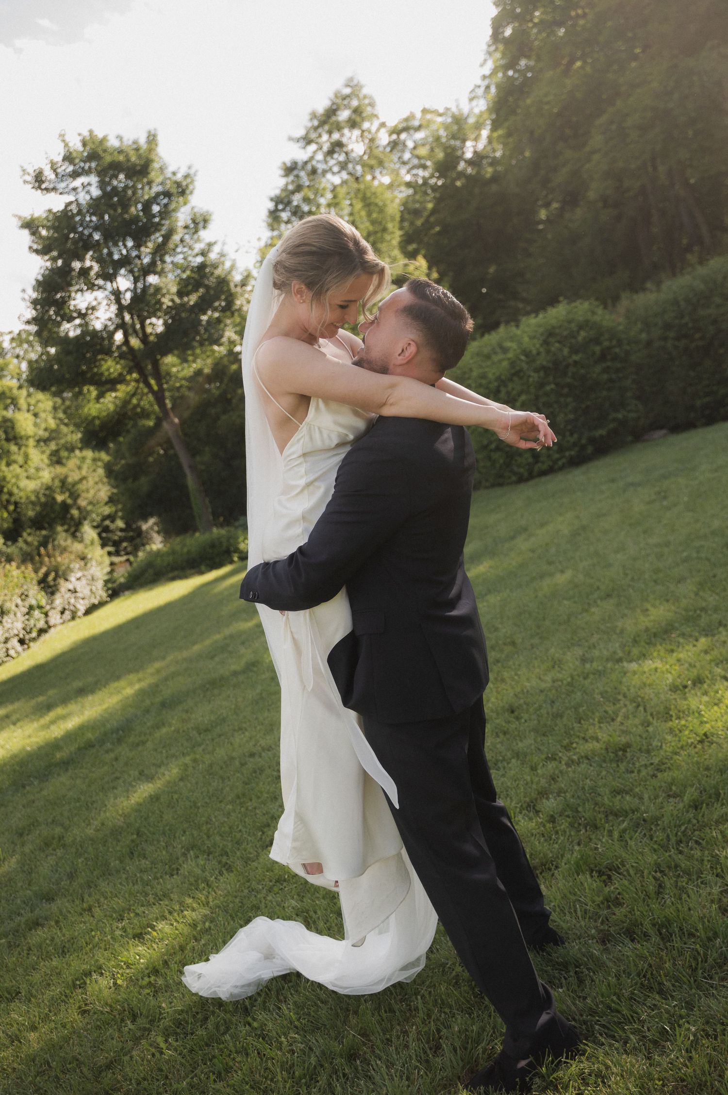 Groom lifting bride in white wedding dress outdoors on lush green lawn surrounded by trees in golden sunlight.