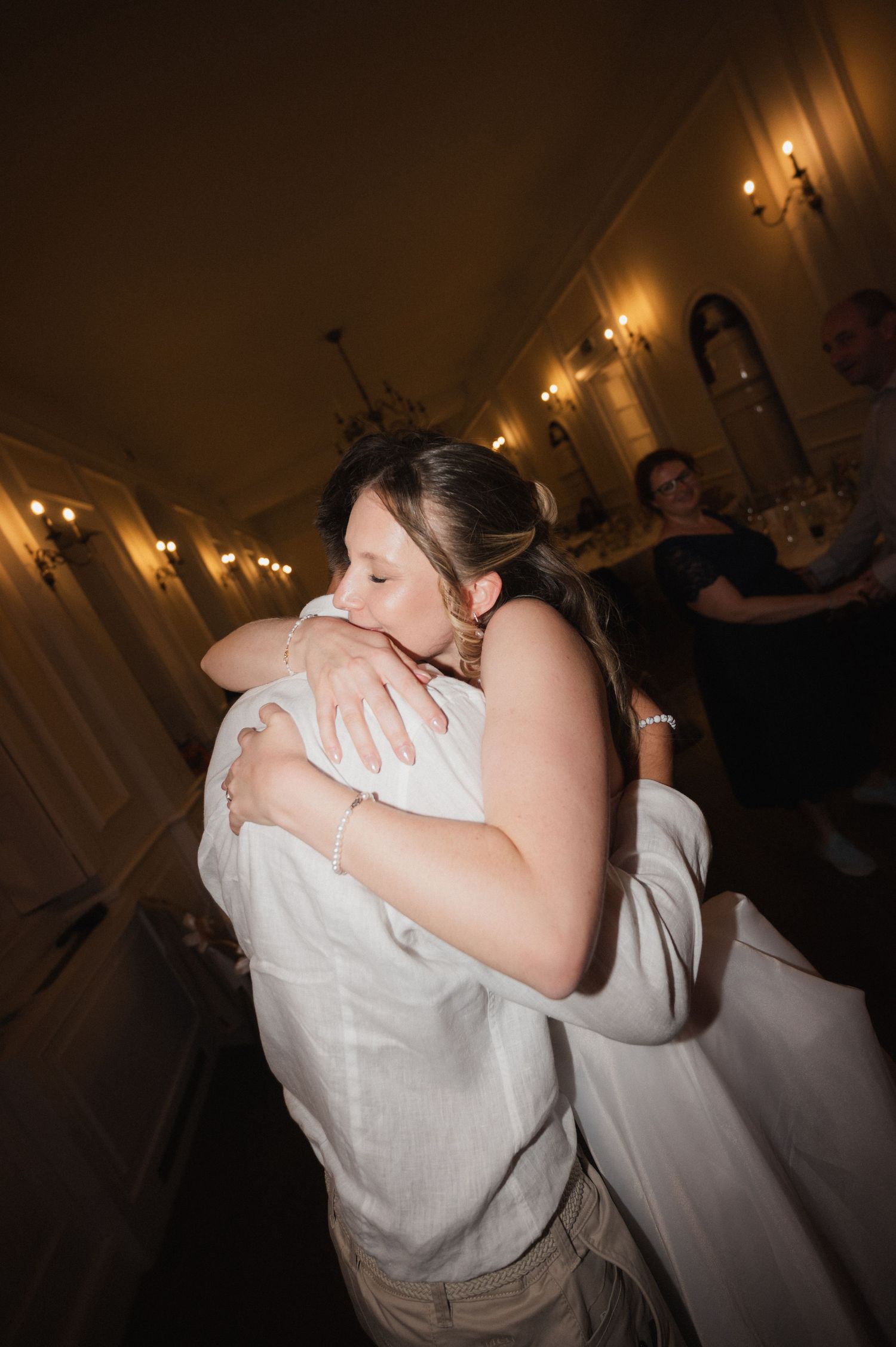 Bride in white dress sharing emotional hug with guest during wedding reception in warmly lit venue.
