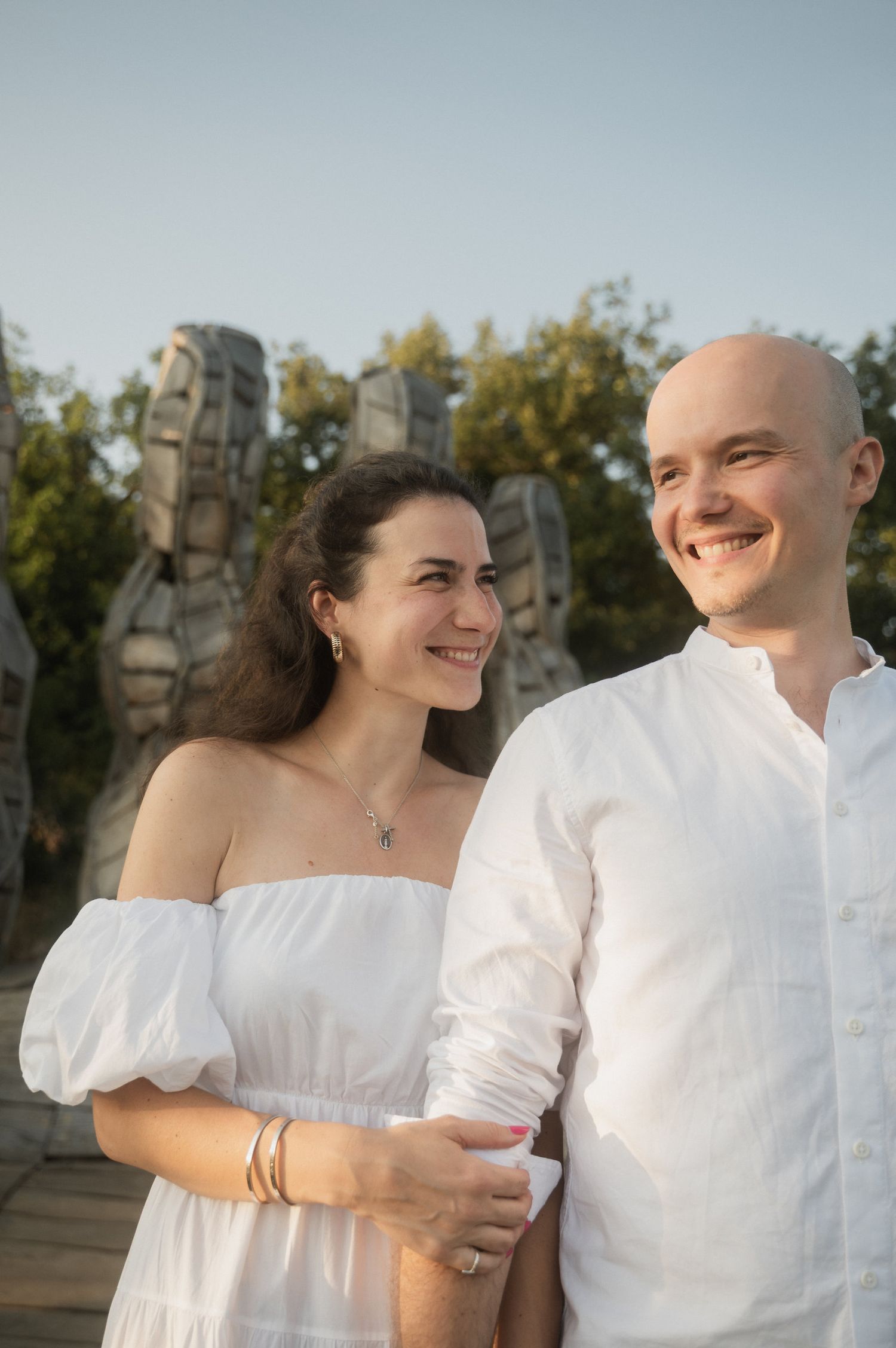 A couple in white clothing smiles together outdoors with stone sculptures visible in the background.