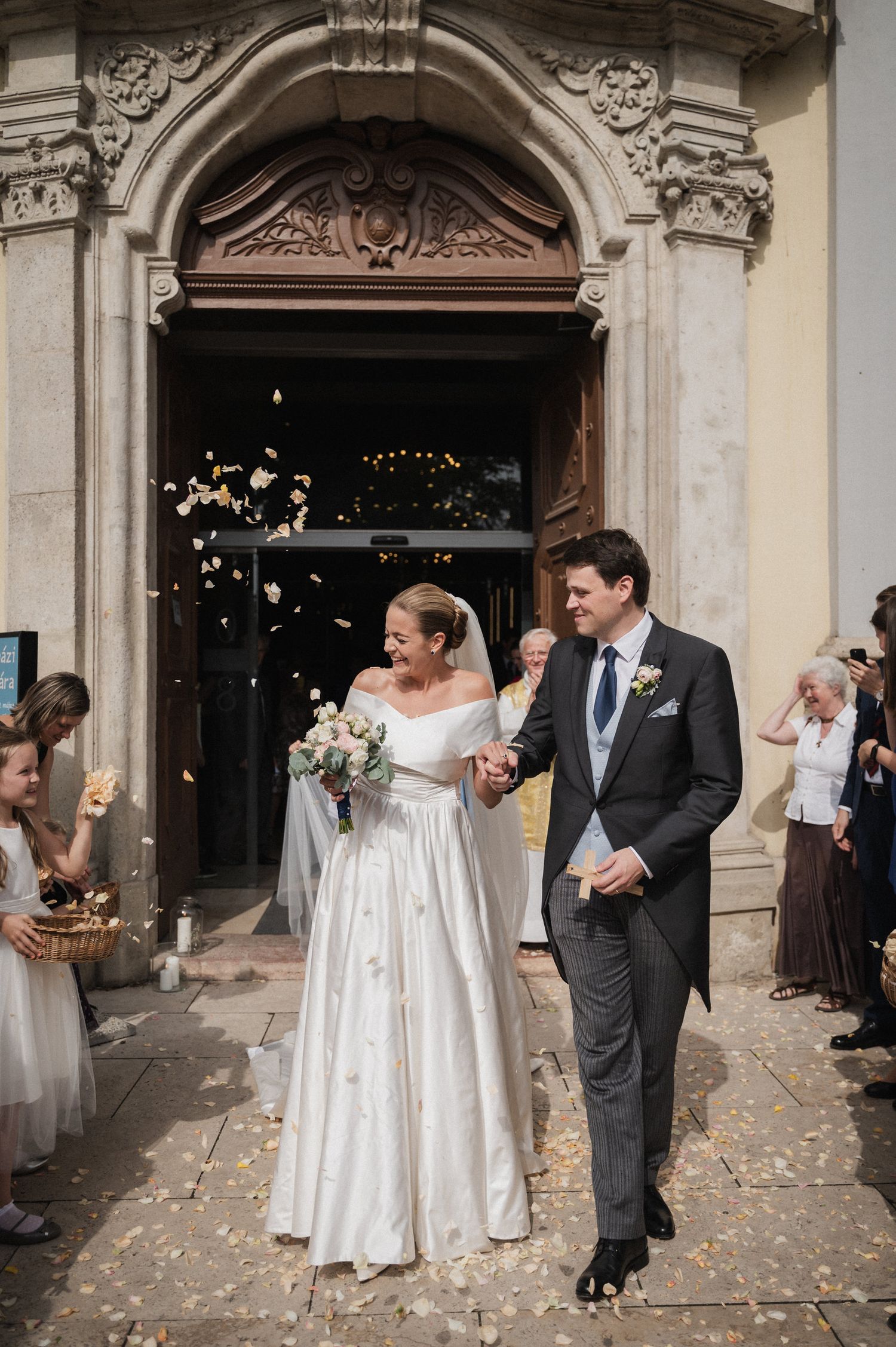 A couple exits a historic church building with ornate stone archway while guests shower them with flower petals.