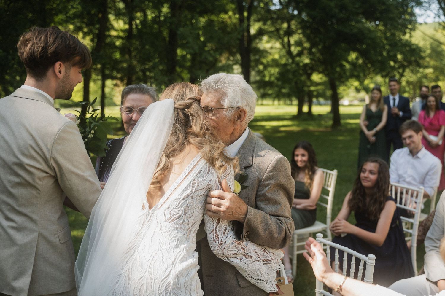 A tender moment at an outdoor wedding ceremony with guests seated on white chairs in a lush garden setting.