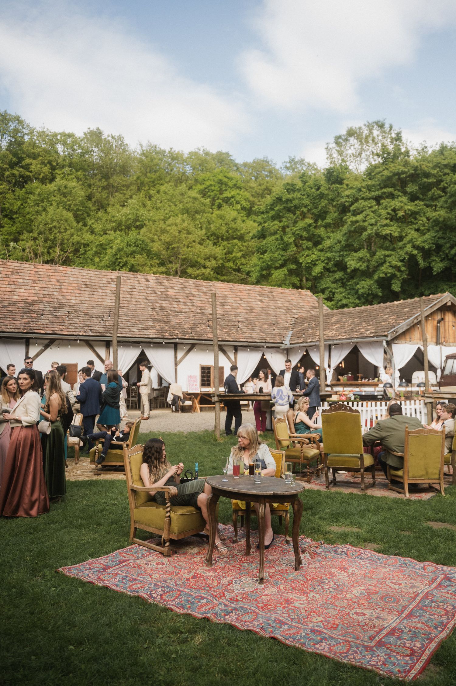 Outdoor event setup with vintage furniture and oriental rugs arranged on grass near rustic buildings.